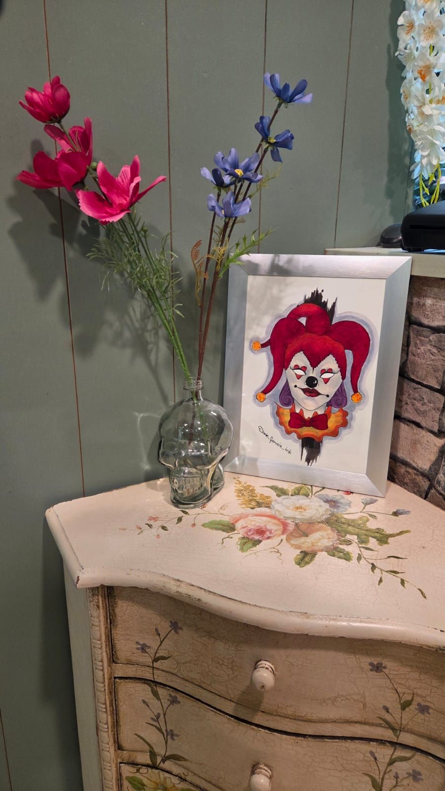 A framed jester illustration, flowers in a skull vase, on a floral-painted cabinet.
