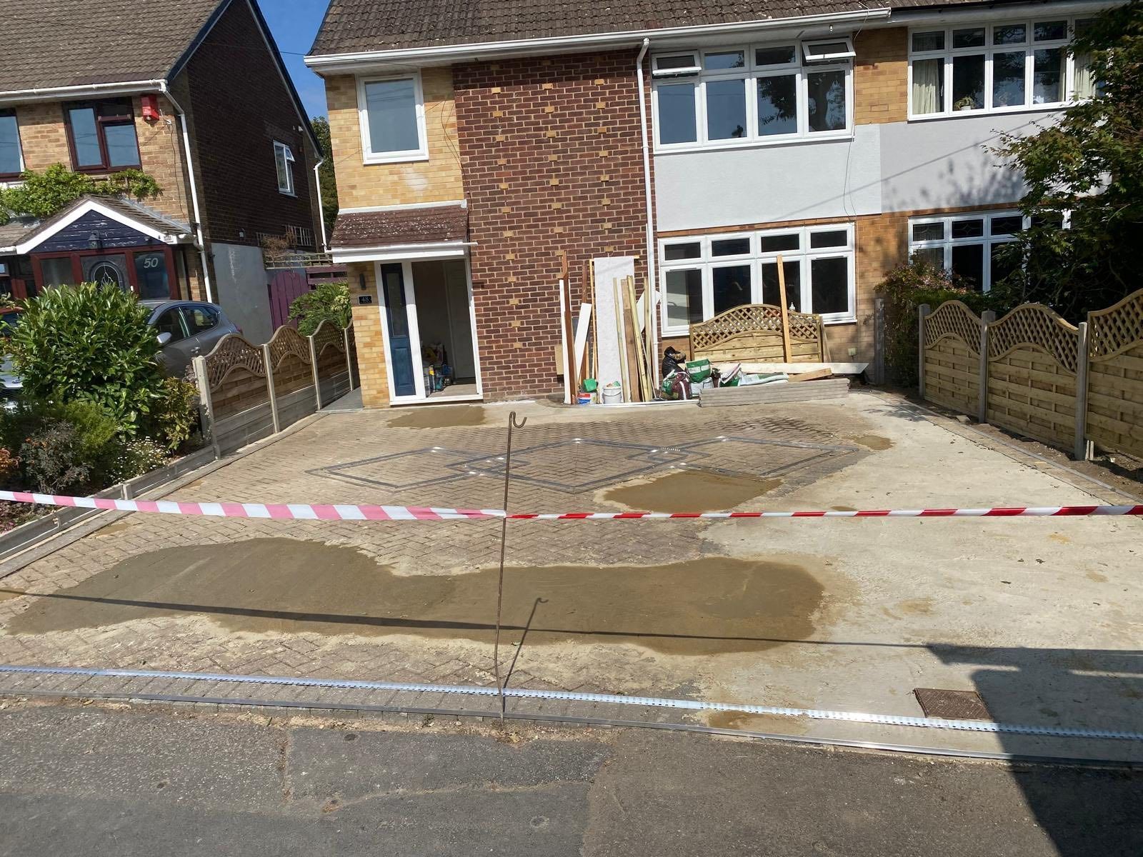 Driveway under construction in front of a two-story house; areas of new concrete, wood, and caution tape.
