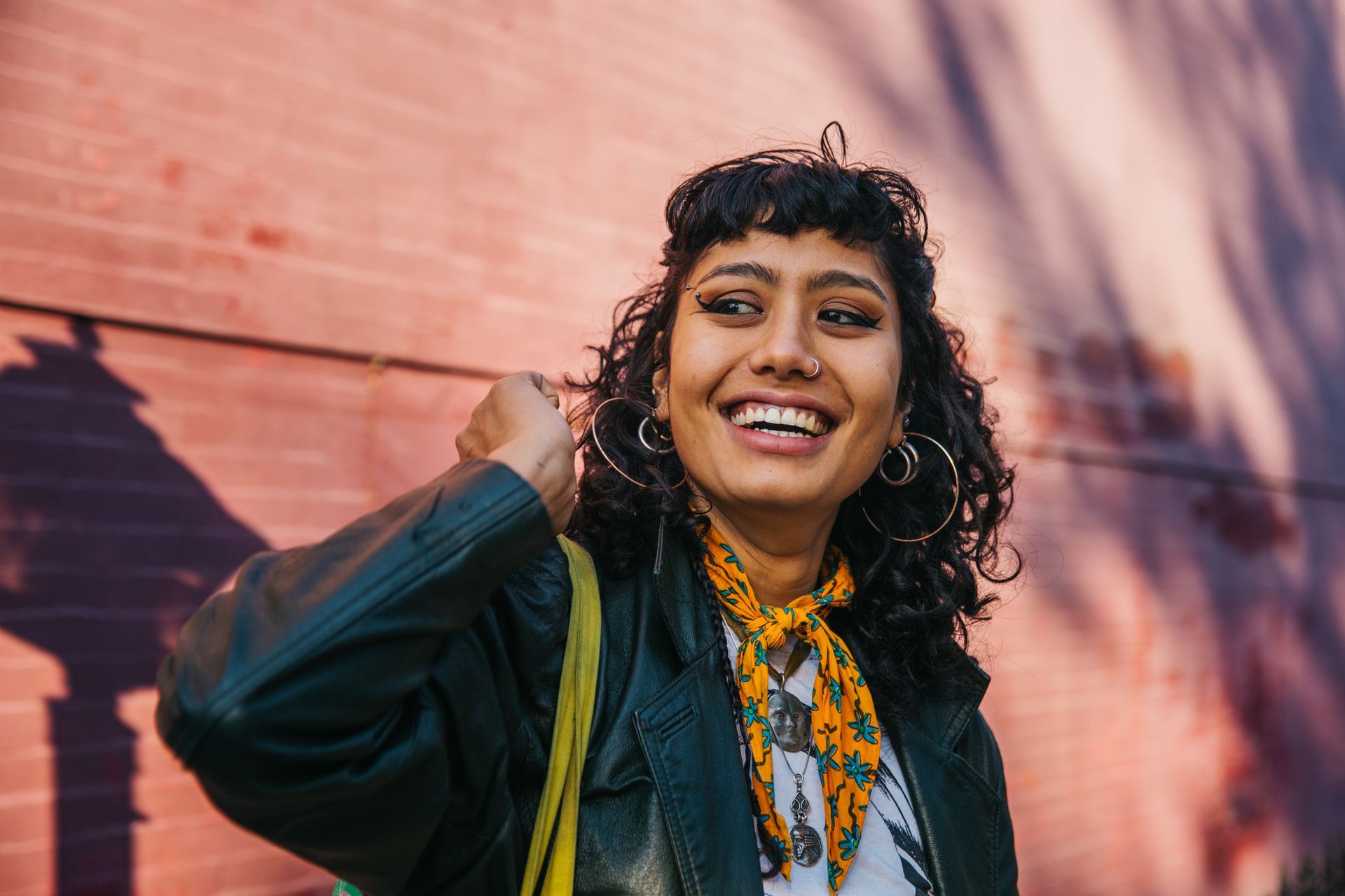A woman wearing a leather jacket and a scarf is smiling in front of a brick wall.