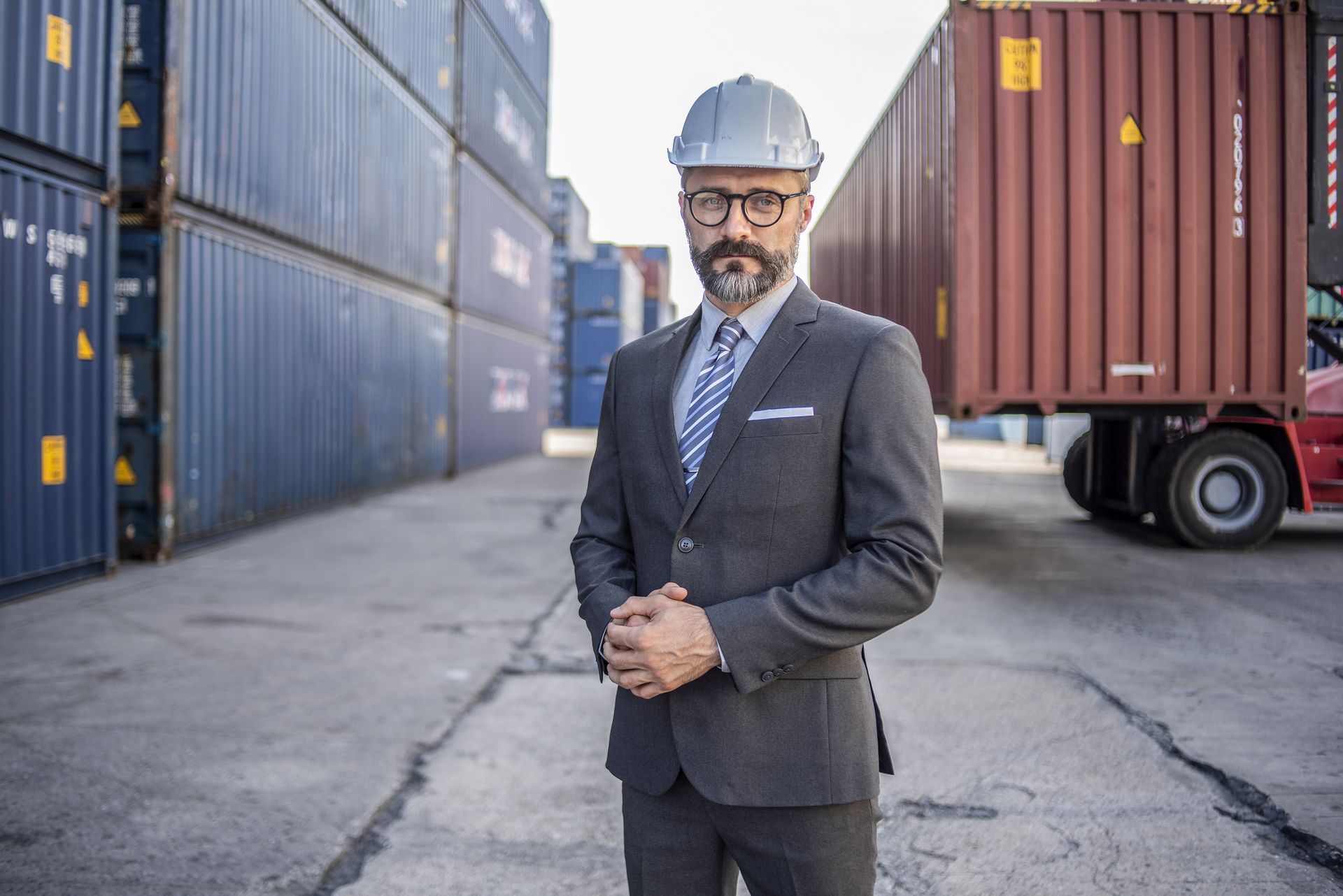 A man in a suit and hard hat is standing in front of shipping containers.