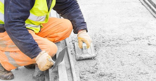 A construction worker is kneeling down and using a trowel to spread concrete.
