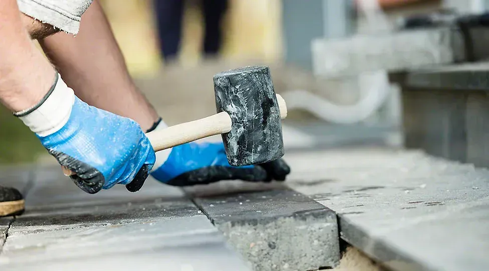 A person is using a hammer to break a brick on a sidewalk.
