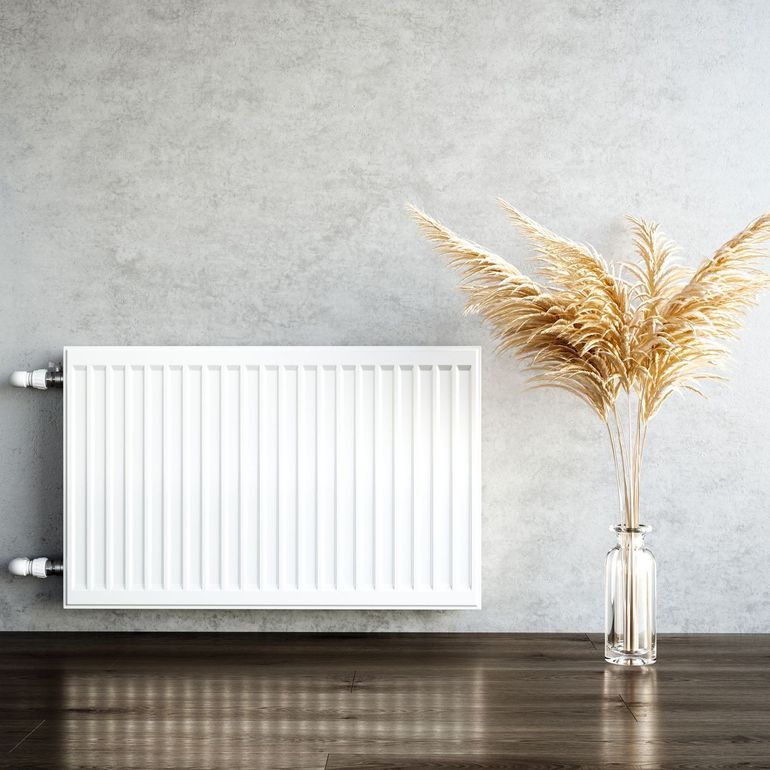 Ein weißer Heizkörper steht auf einem Holzboden neben einer Vase mit getrockneten Blumen.