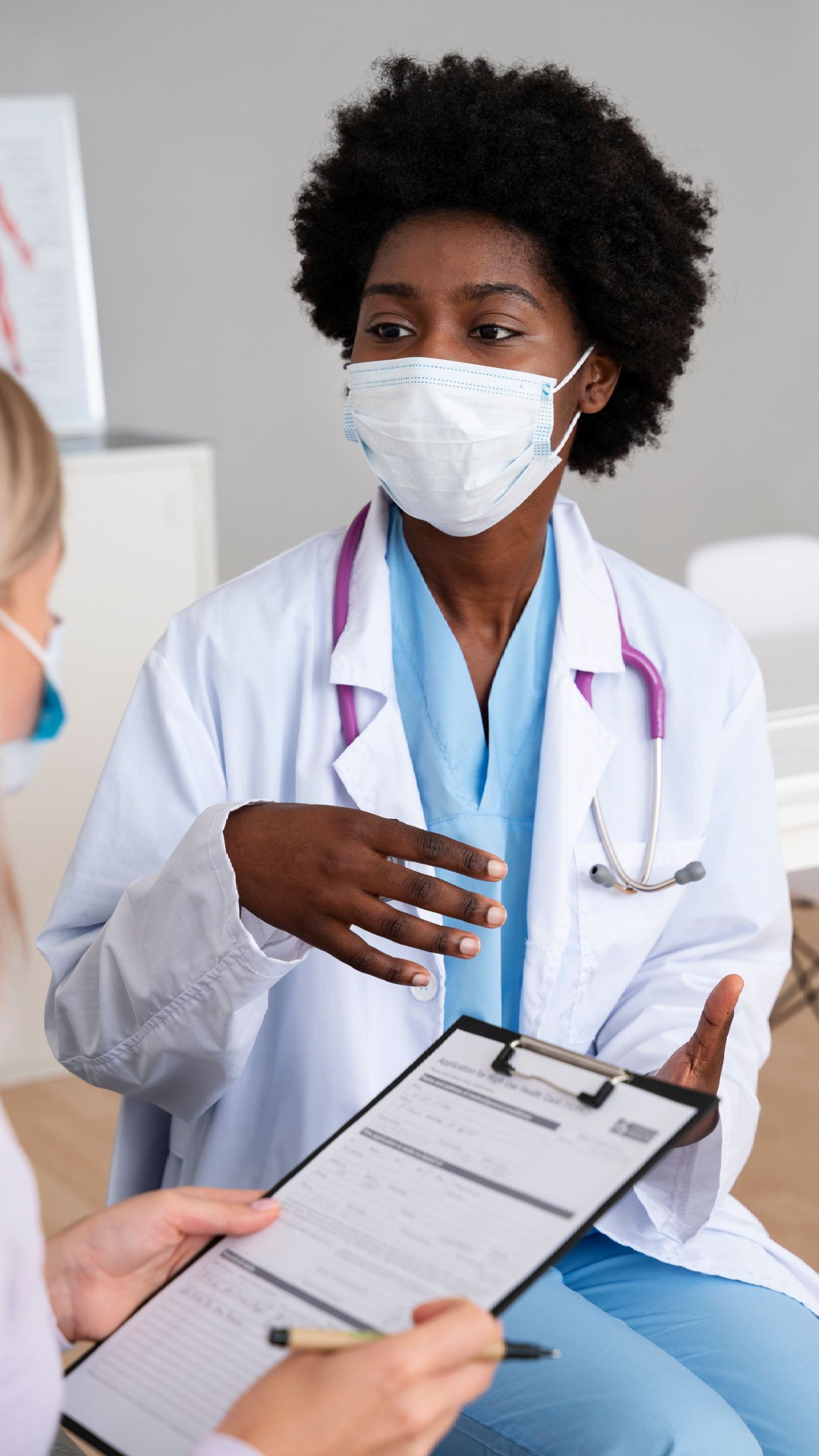 a doctor wearing a mask is talking to a patient while holding a clipboard .