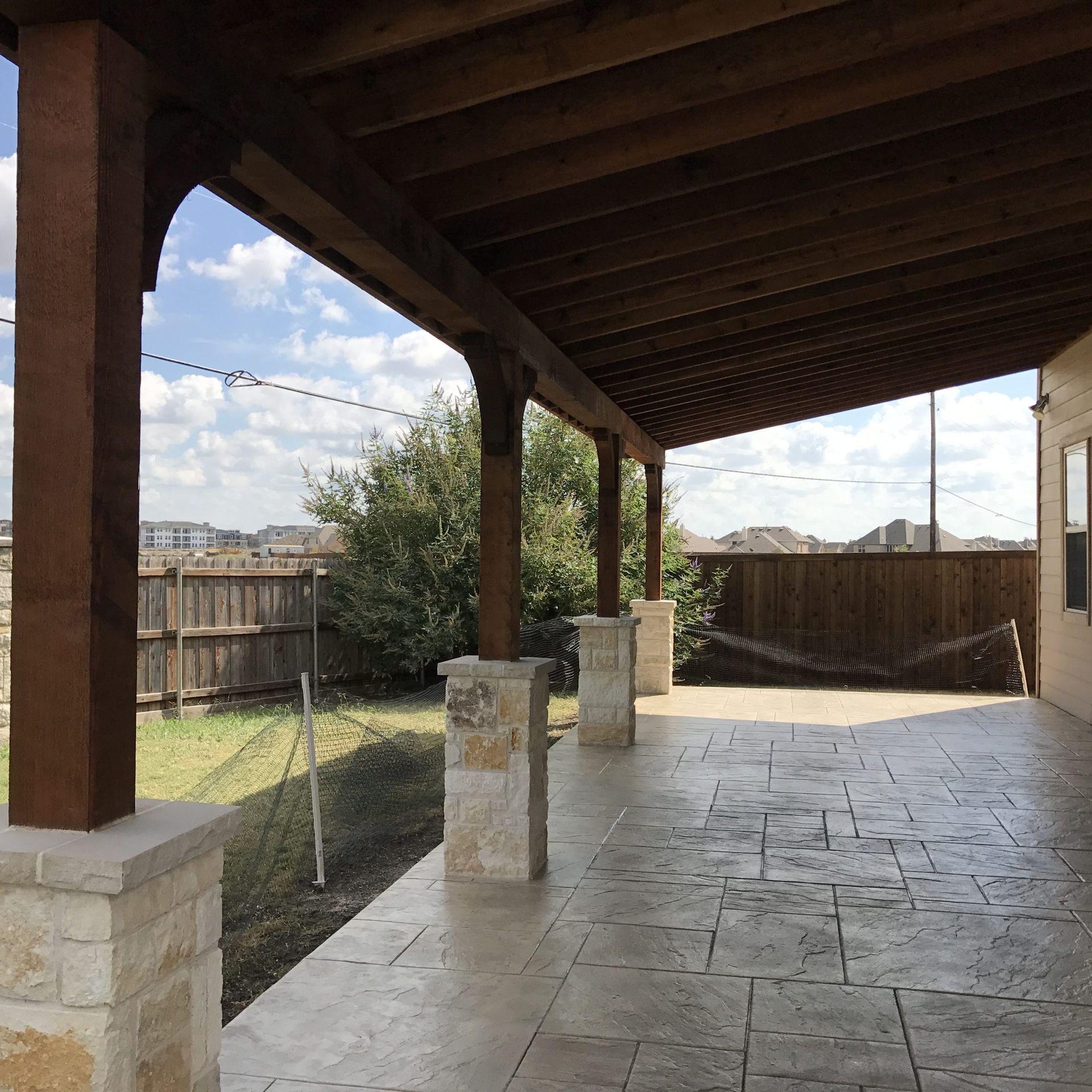 Covered patio with stone columns, dark wood beams, and stamped concrete floor. Backyard view.