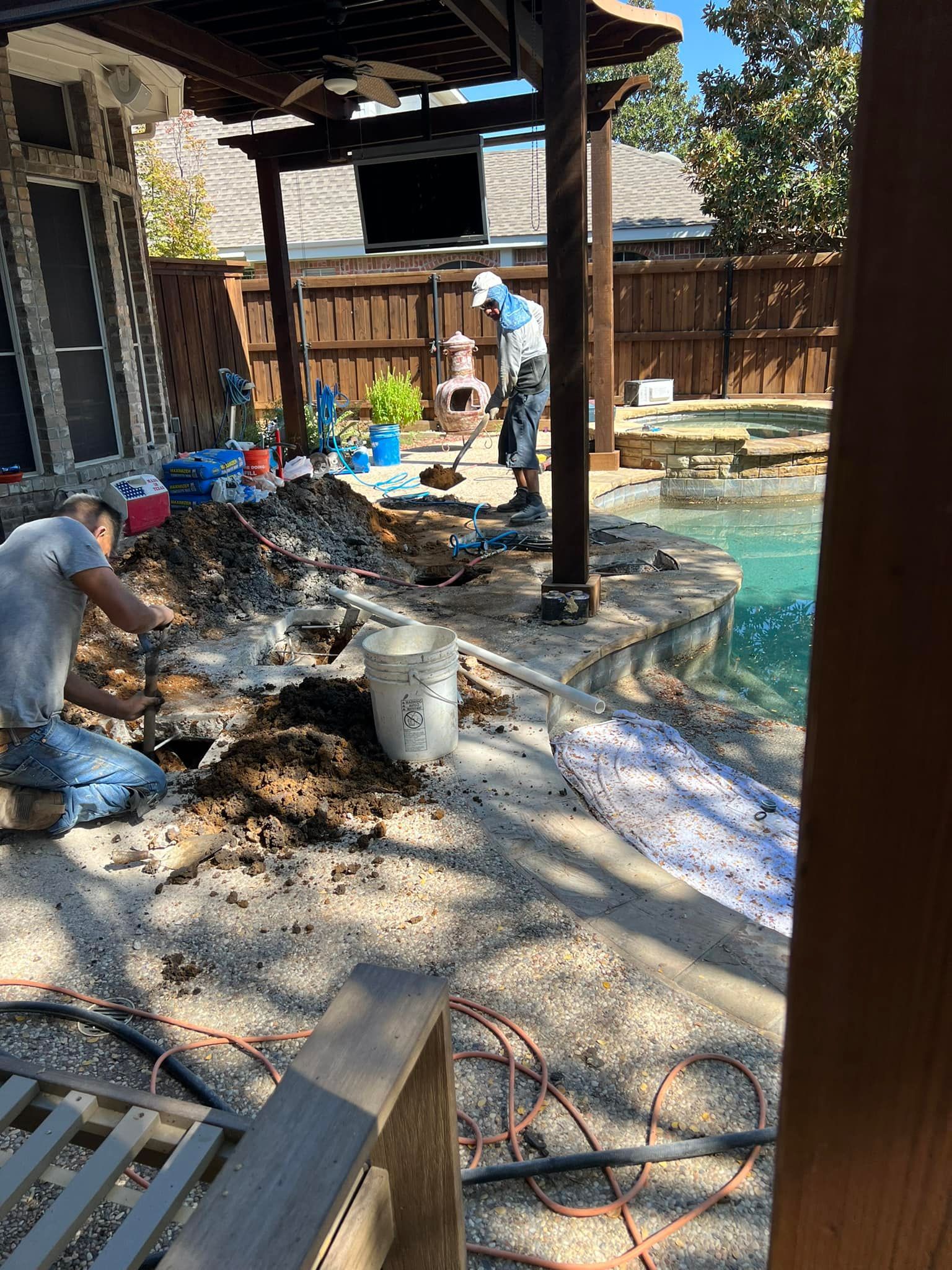 Workers repairing a pool area outdoors. One digs, others work near dirt piles and the pool.