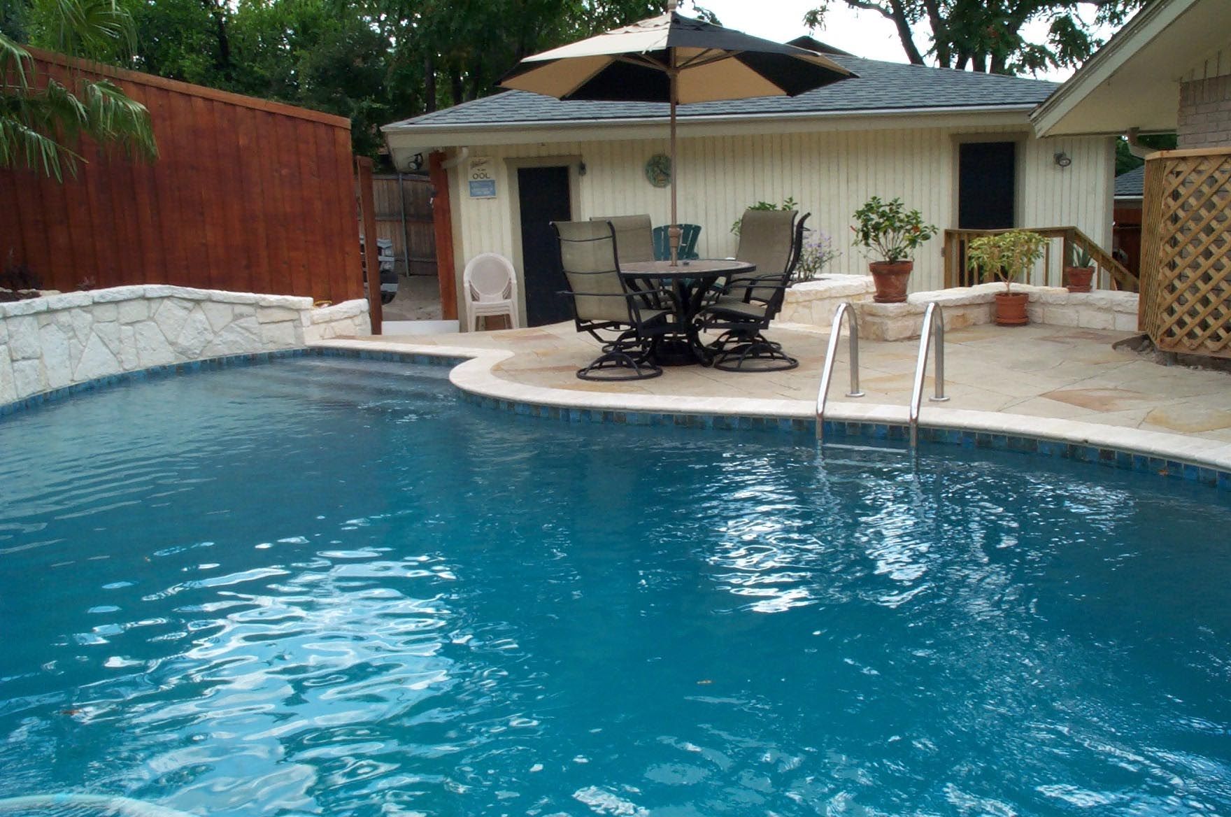 Swimming pool with a patio area, table, and building in the background.