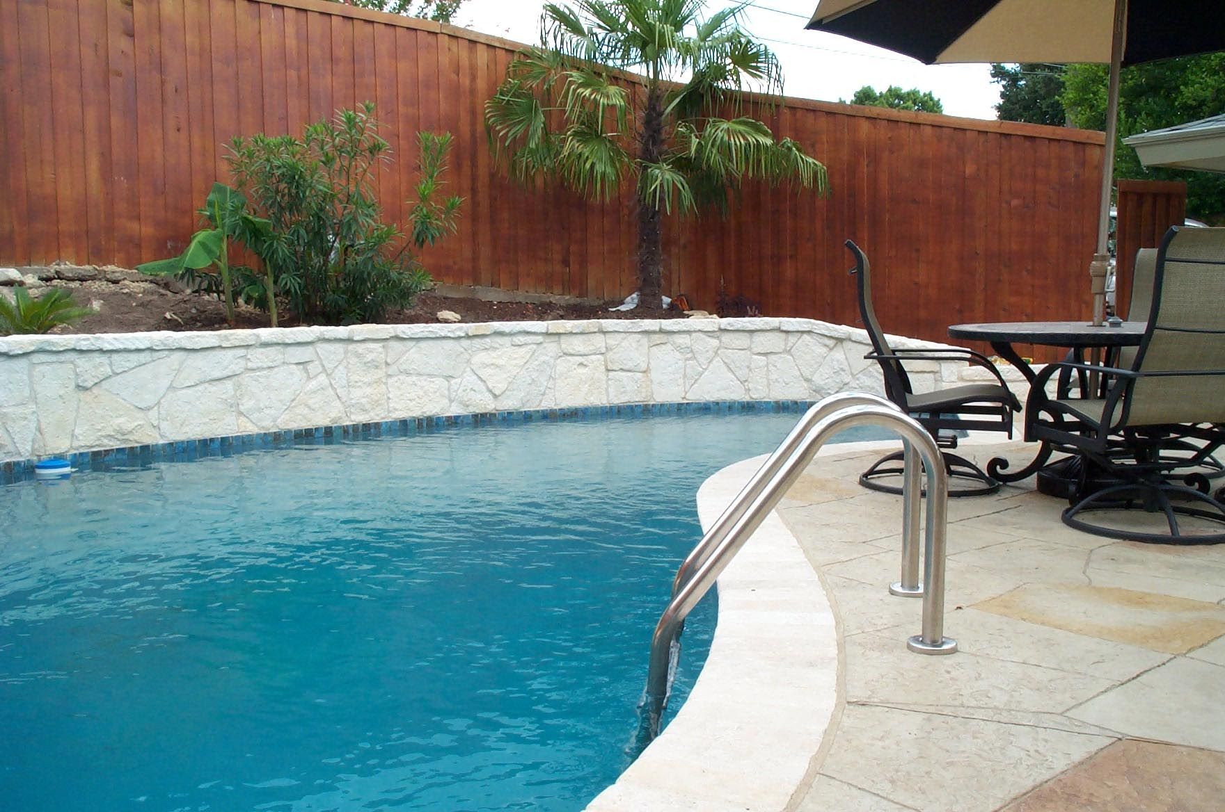 Pool with a stone border, next to patio furniture and a wooden fence.