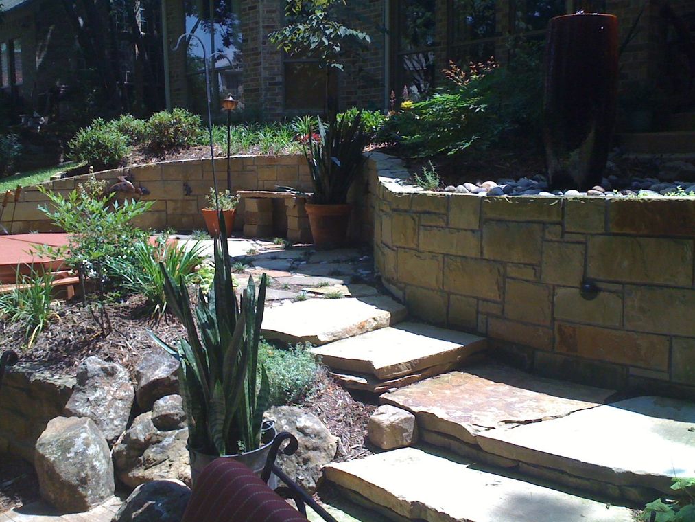 Stone steps leading through a terraced garden with a water feature. Sunny, outdoor setting.
