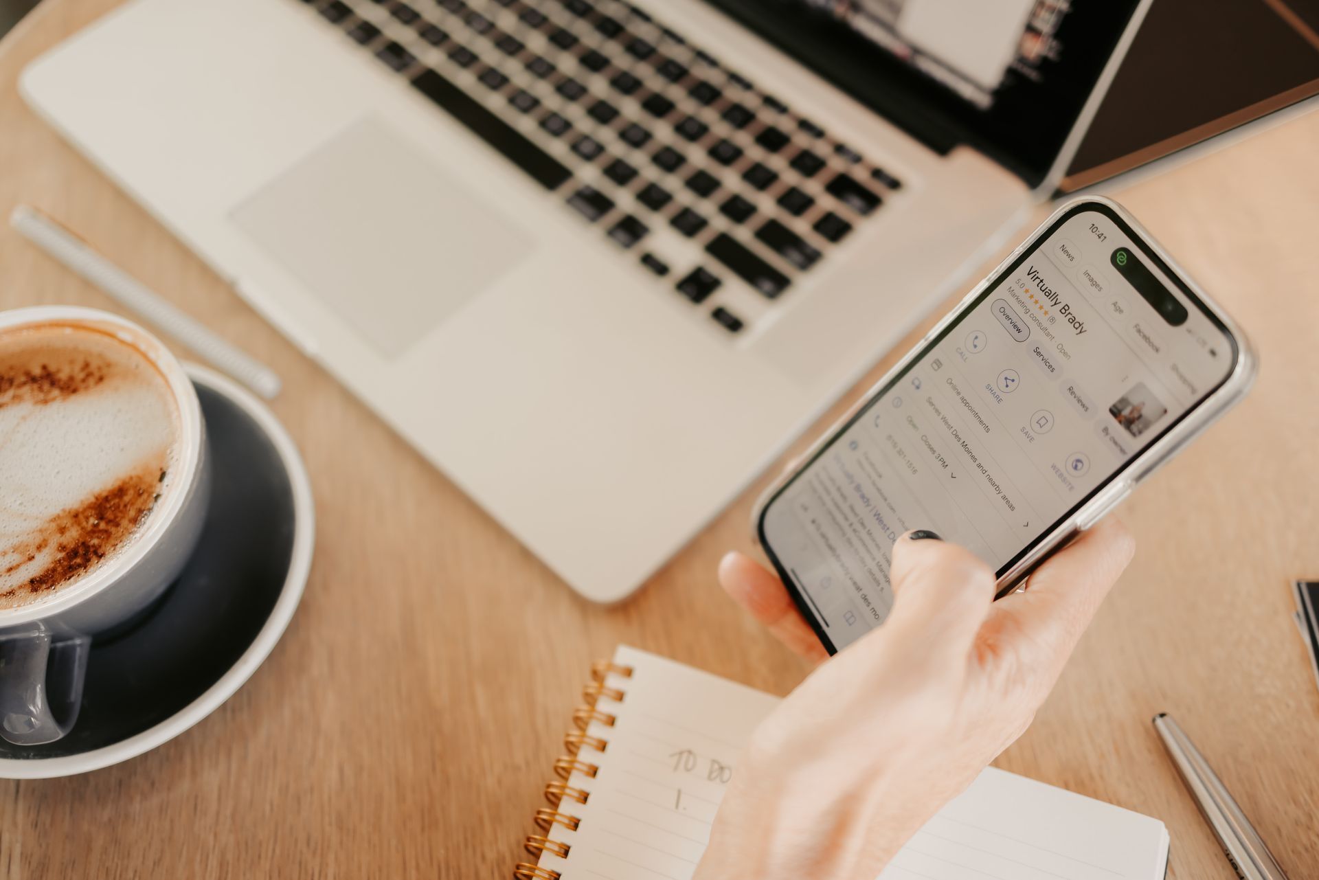 A person is using a cell phone in front of a laptop and a cup of coffee.