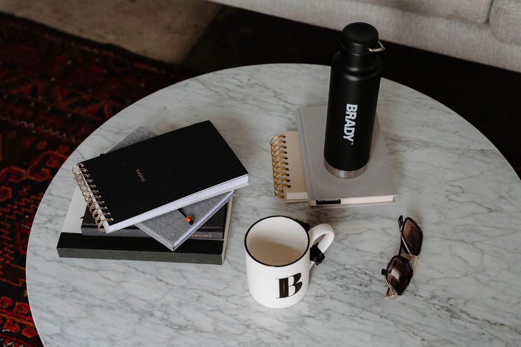 A coffee table with a cup , notebooks , a water bottle and sunglasses on it.