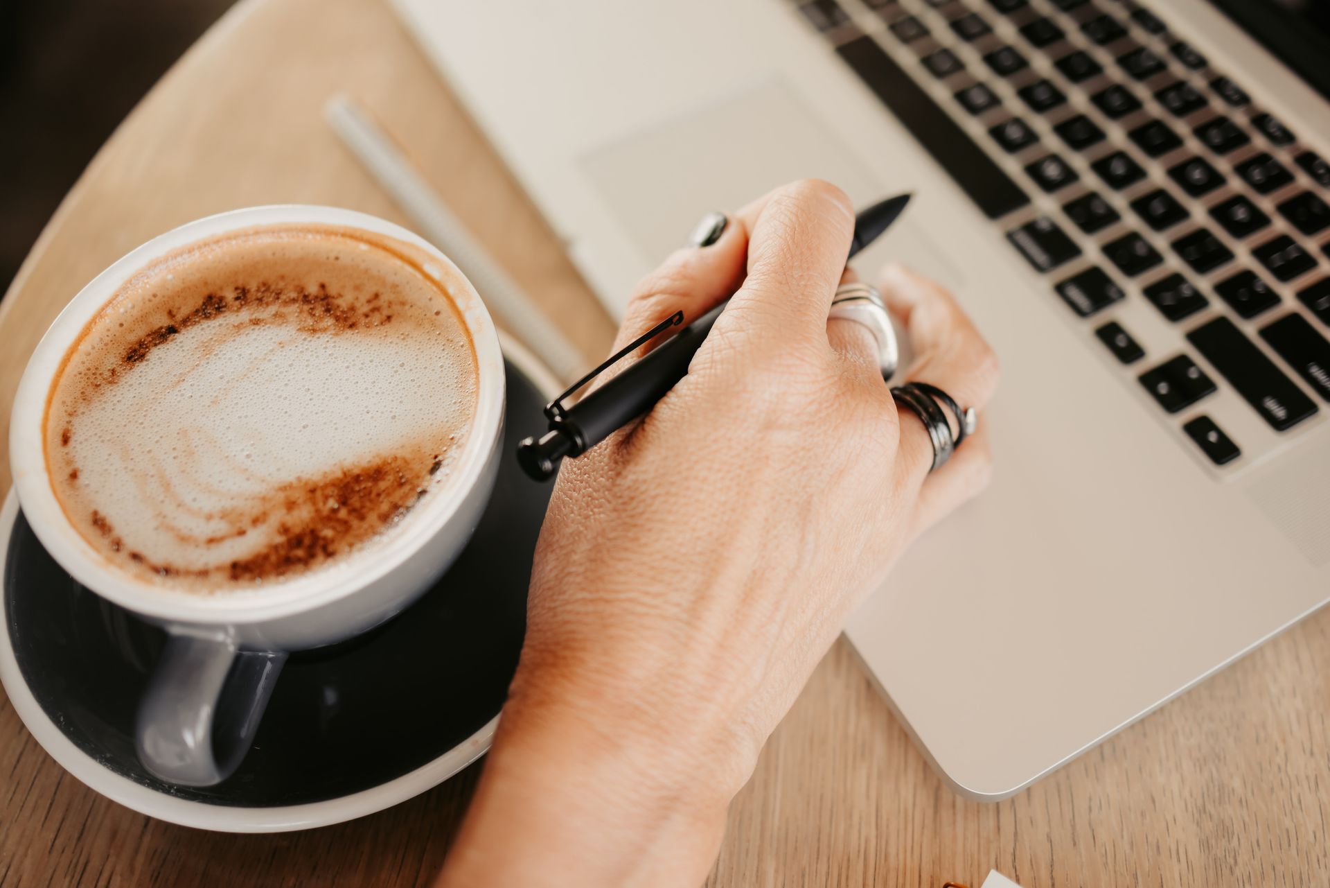 Hand holding a pen beside a laptop and a cup of coffee on a wooden desk