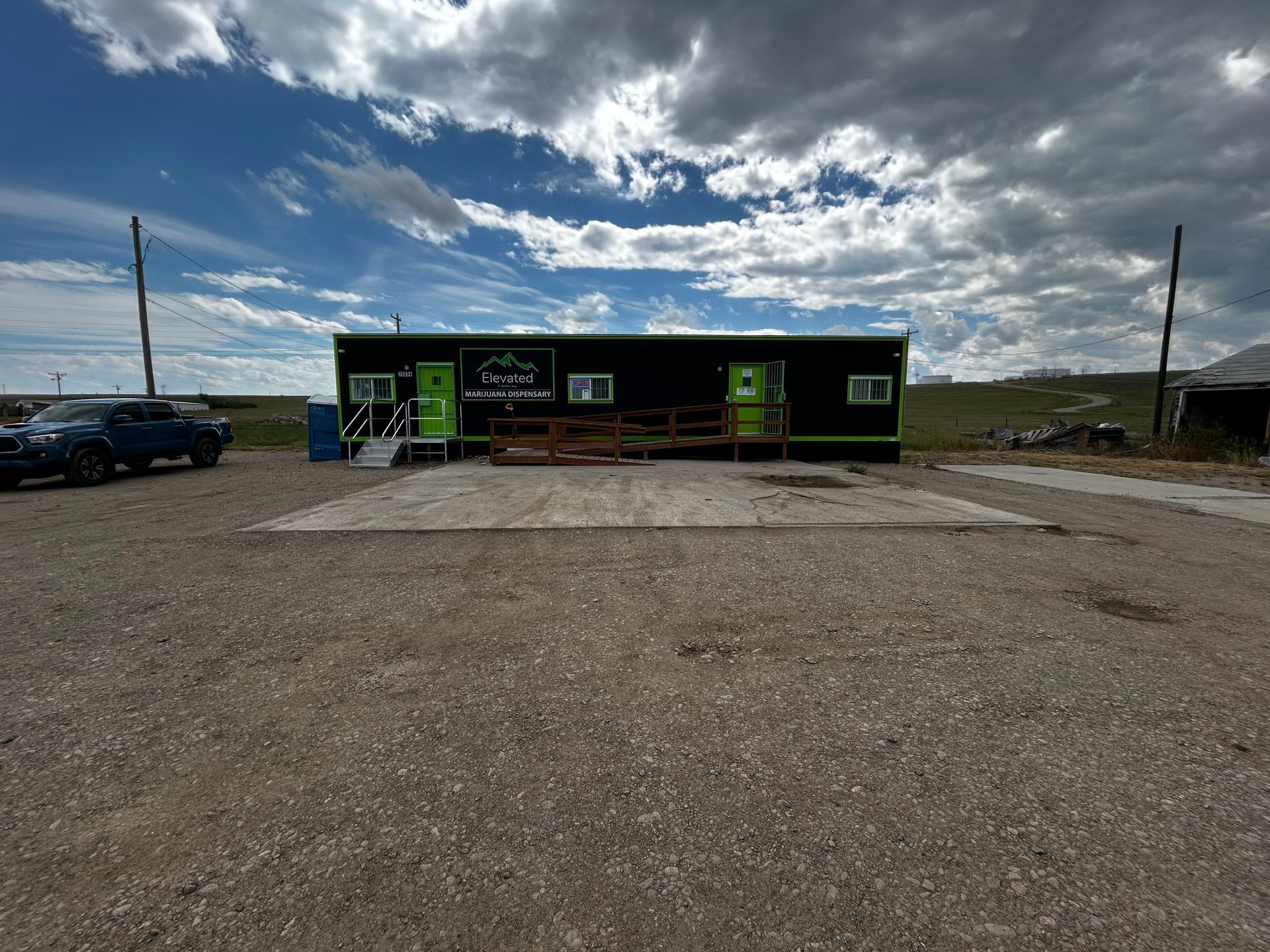 Black building with two green doors and a wooden ramp under a cloudy sky.