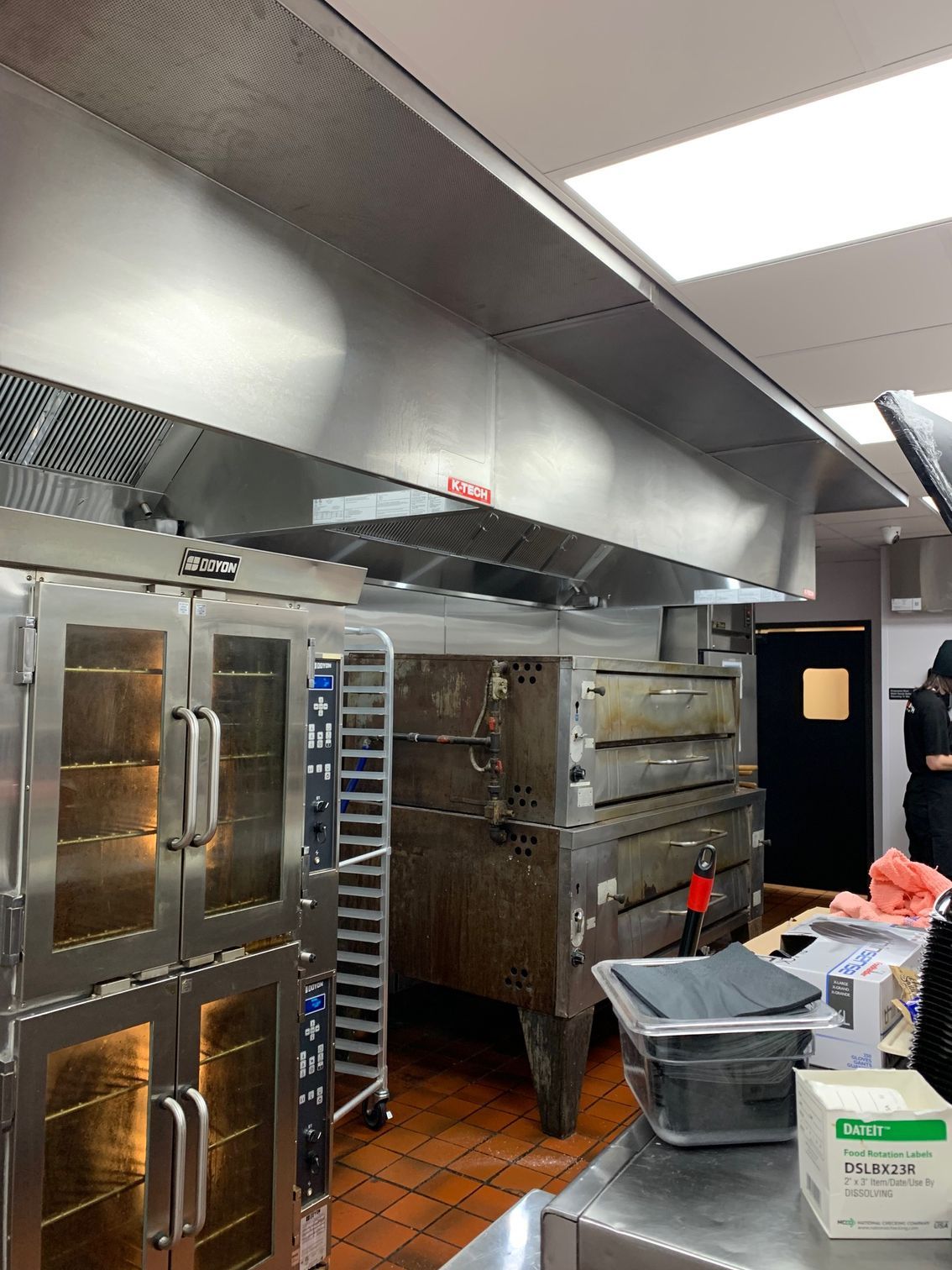 Commercial kitchen with ovens under a stainless steel hood; a person stands near a doorway.