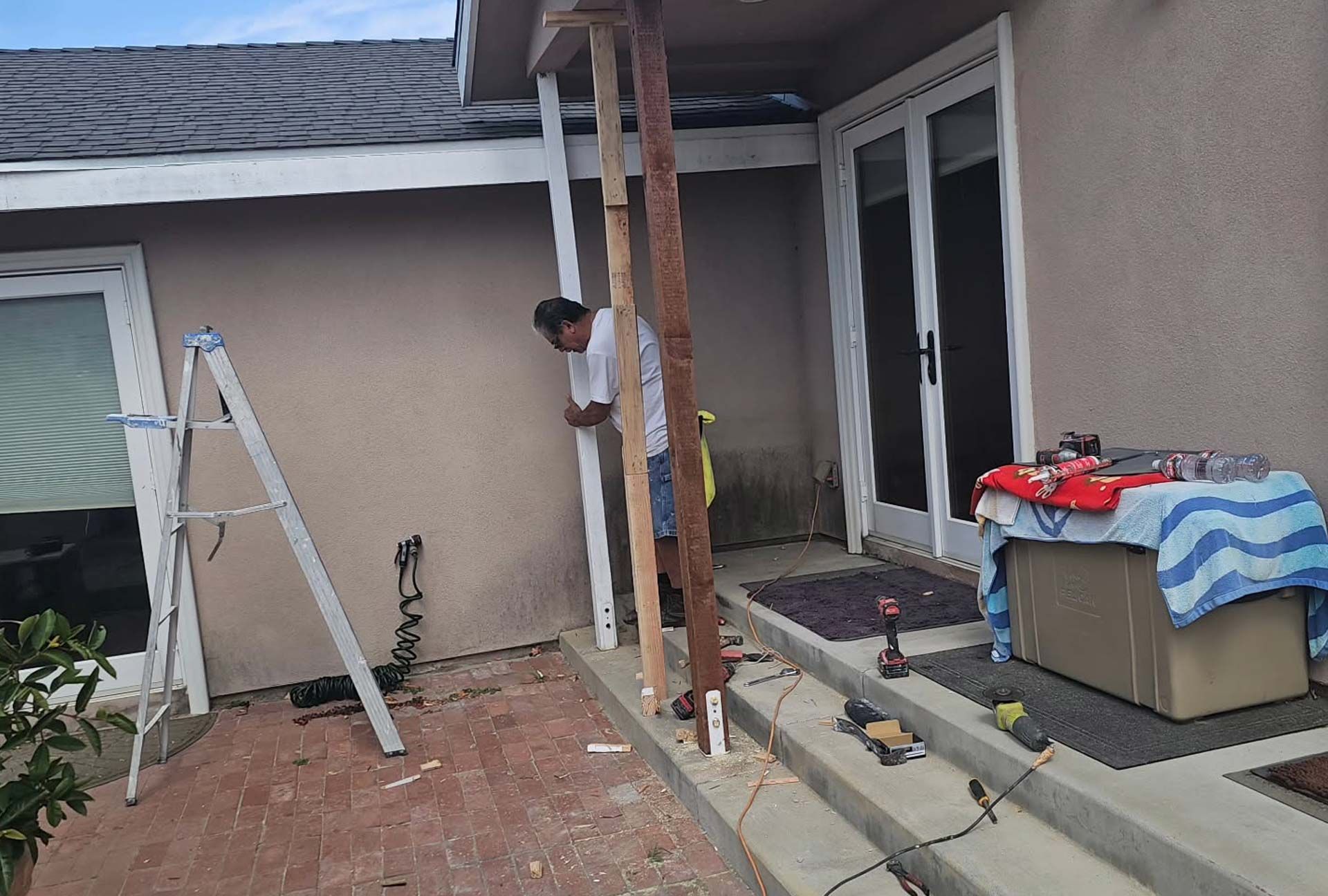 Man working on porch columns outdoors. Ladder beside stucco wall and sliding glass door.