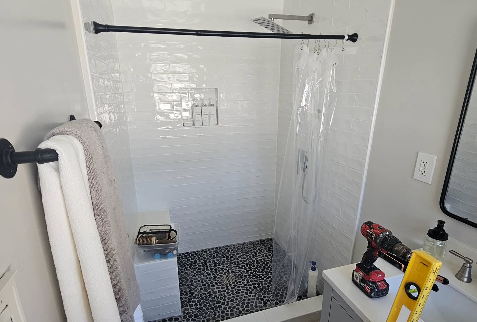 Bathroom with white tiled shower, black pebble floor, shower curtain, towel rack, and vanity with a drill.