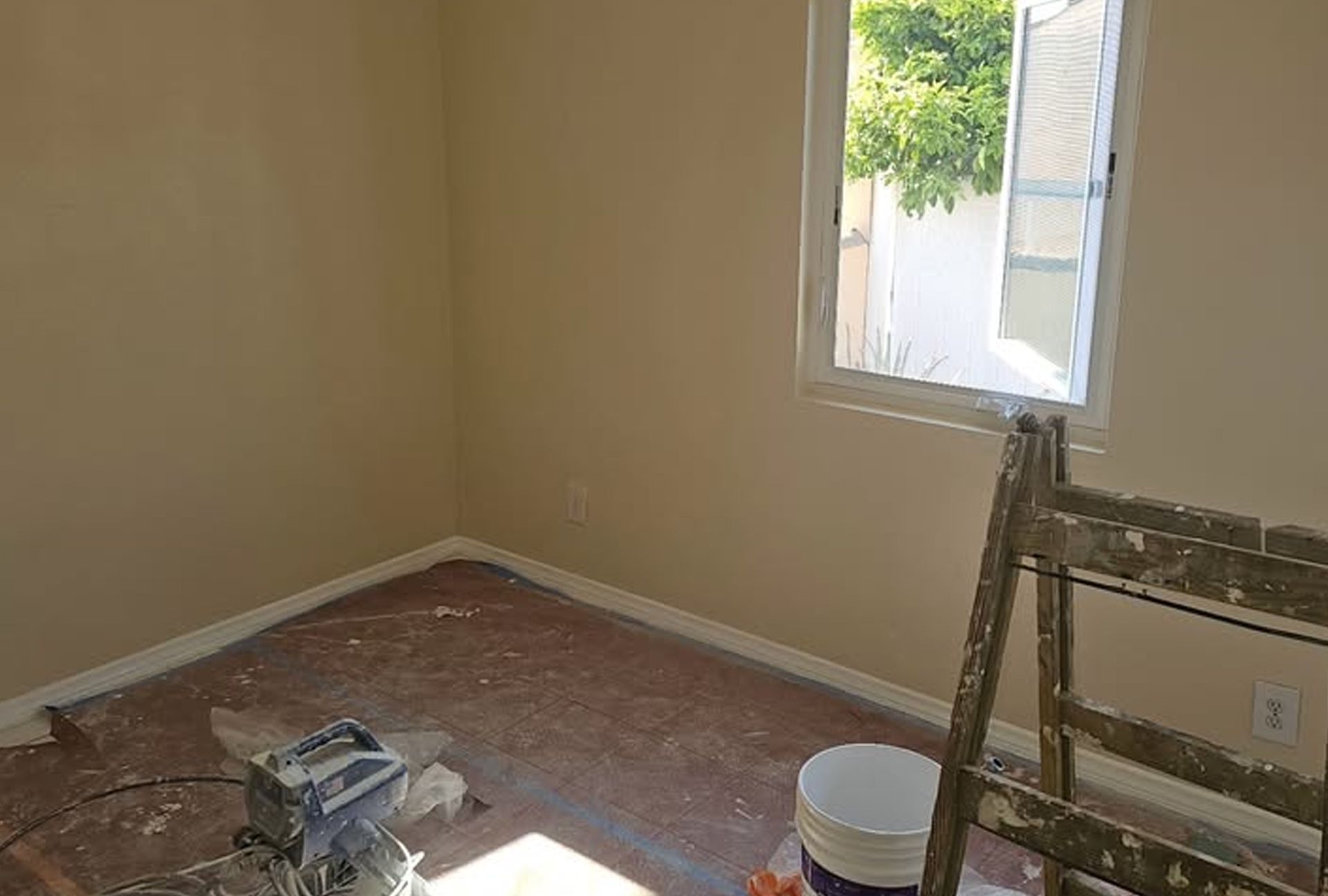 Interior room with tan walls, window, ladder, and paint supplies; floor has protective covering.