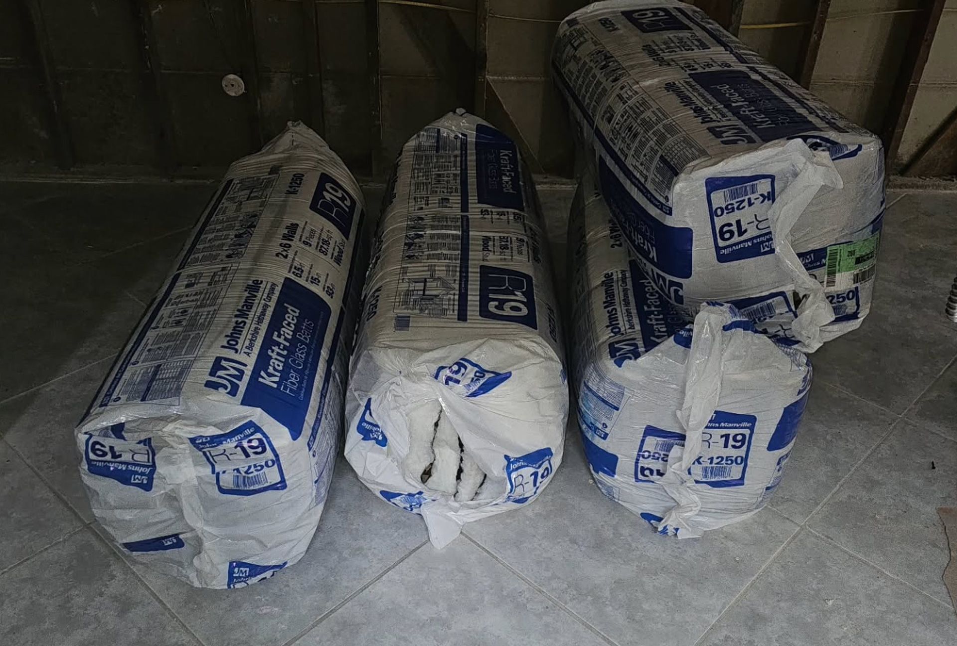 Four bundles of insulation in plastic wrap, stacked on a floor in a room.