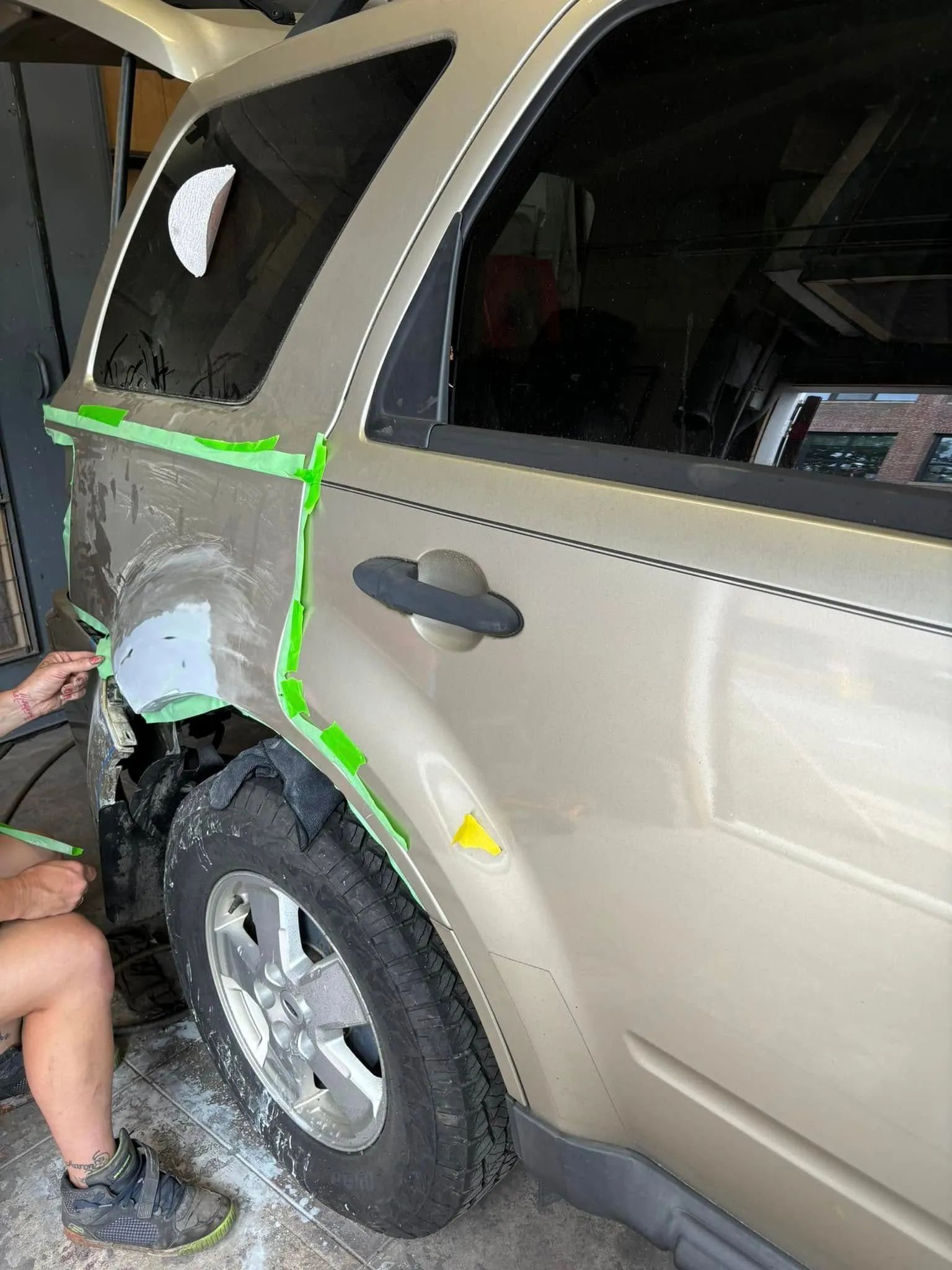 A tan SUV being repaired in a garage; areas taped with green, damaged panel attached. A person's arm and leg are visible.