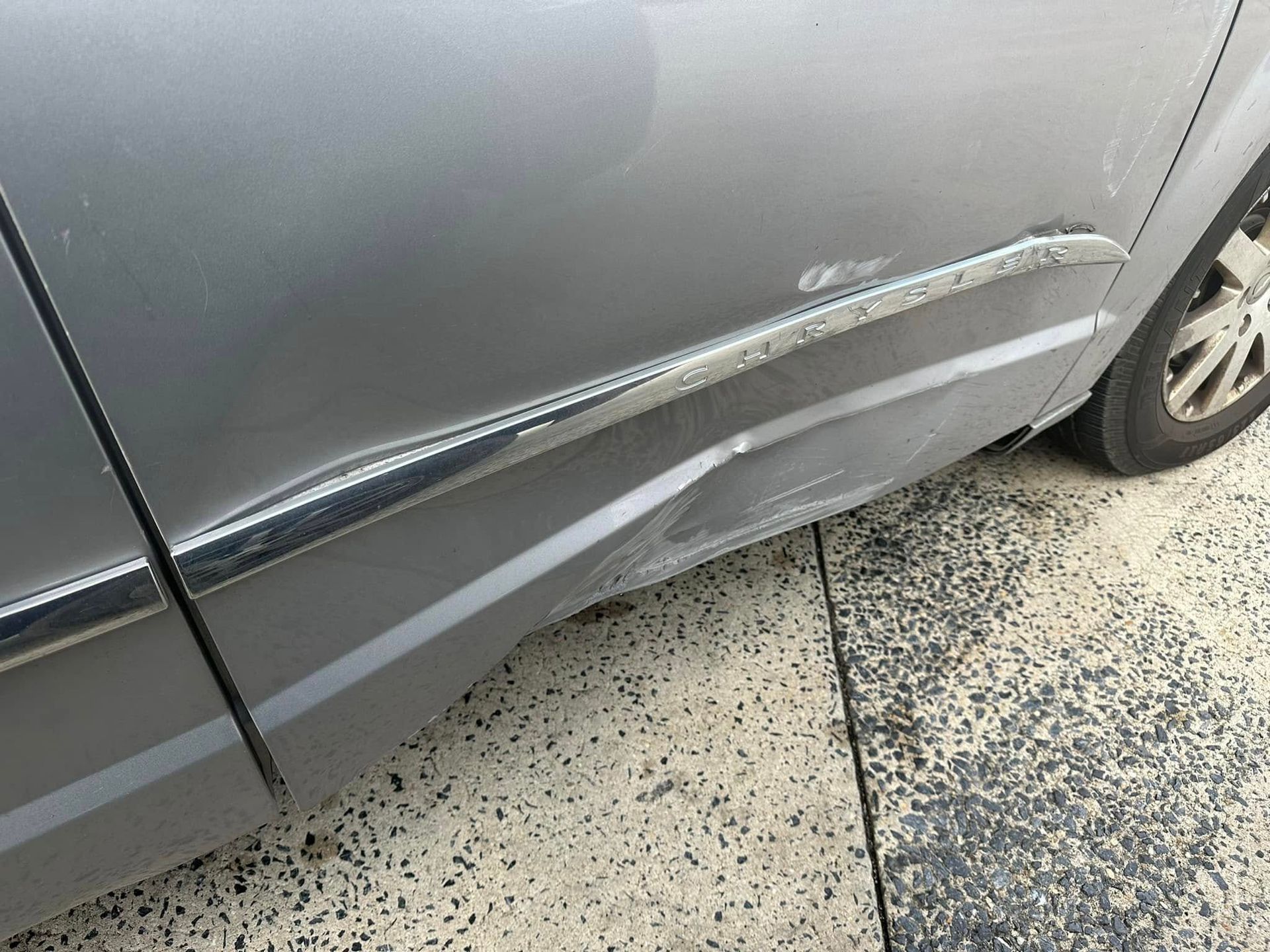 A silver car with a damaged side door is parked on the side of the road.