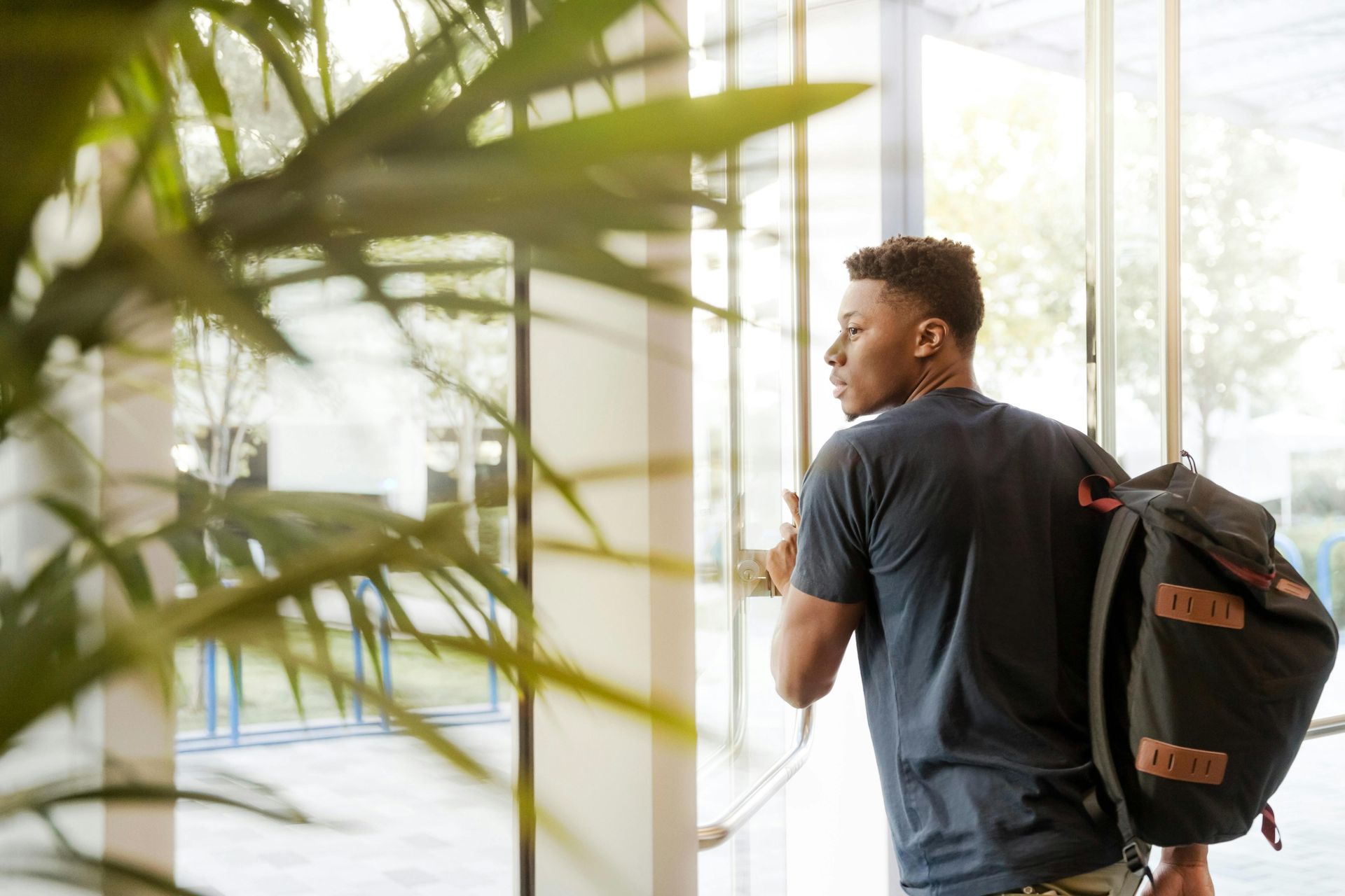 Young Black man with backpack opening a glass door, looking outside.