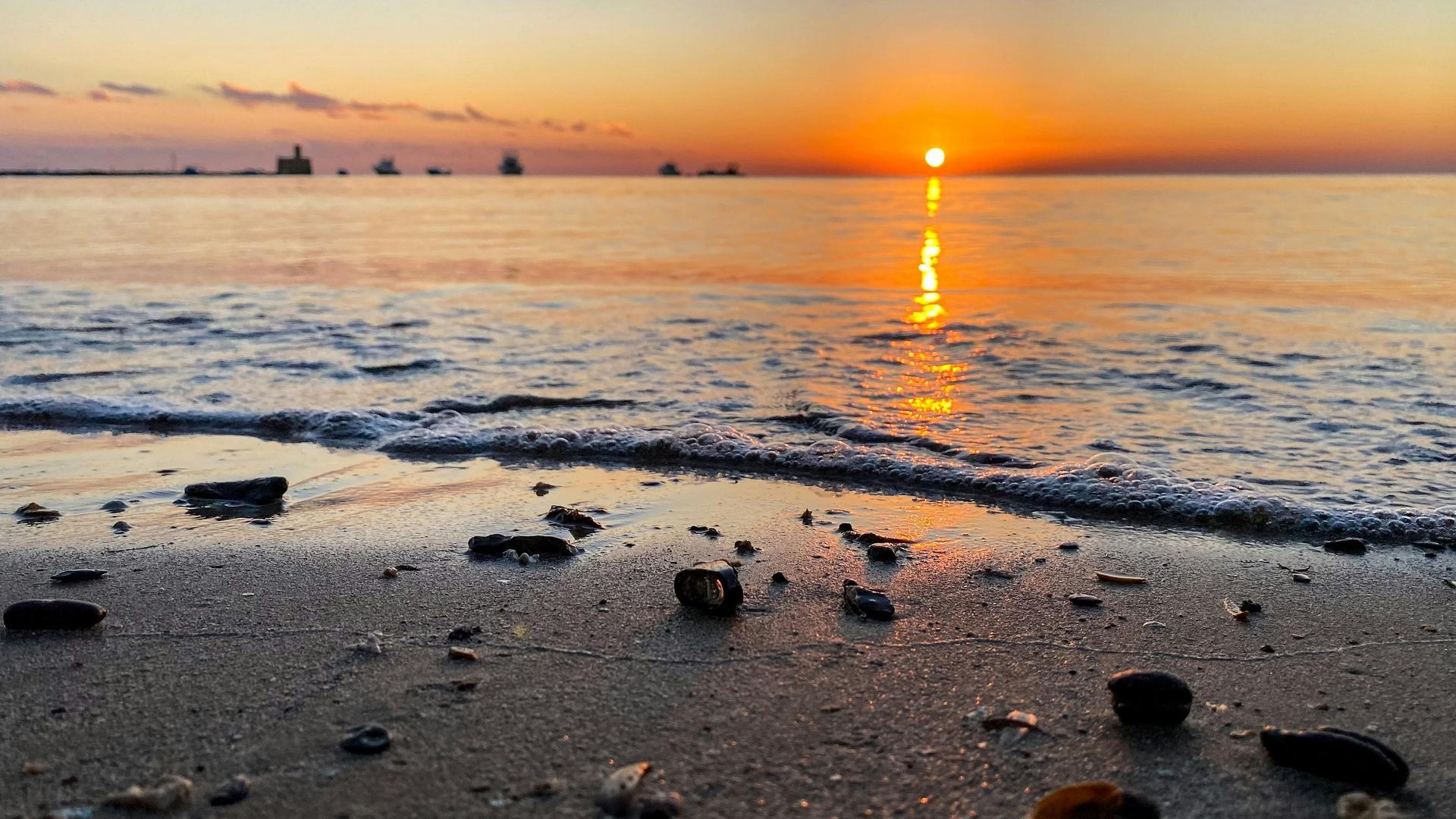 Beach sunset with orange sky reflecting on water; dark silhouettes of ships on the horizon, rocks on the sand.