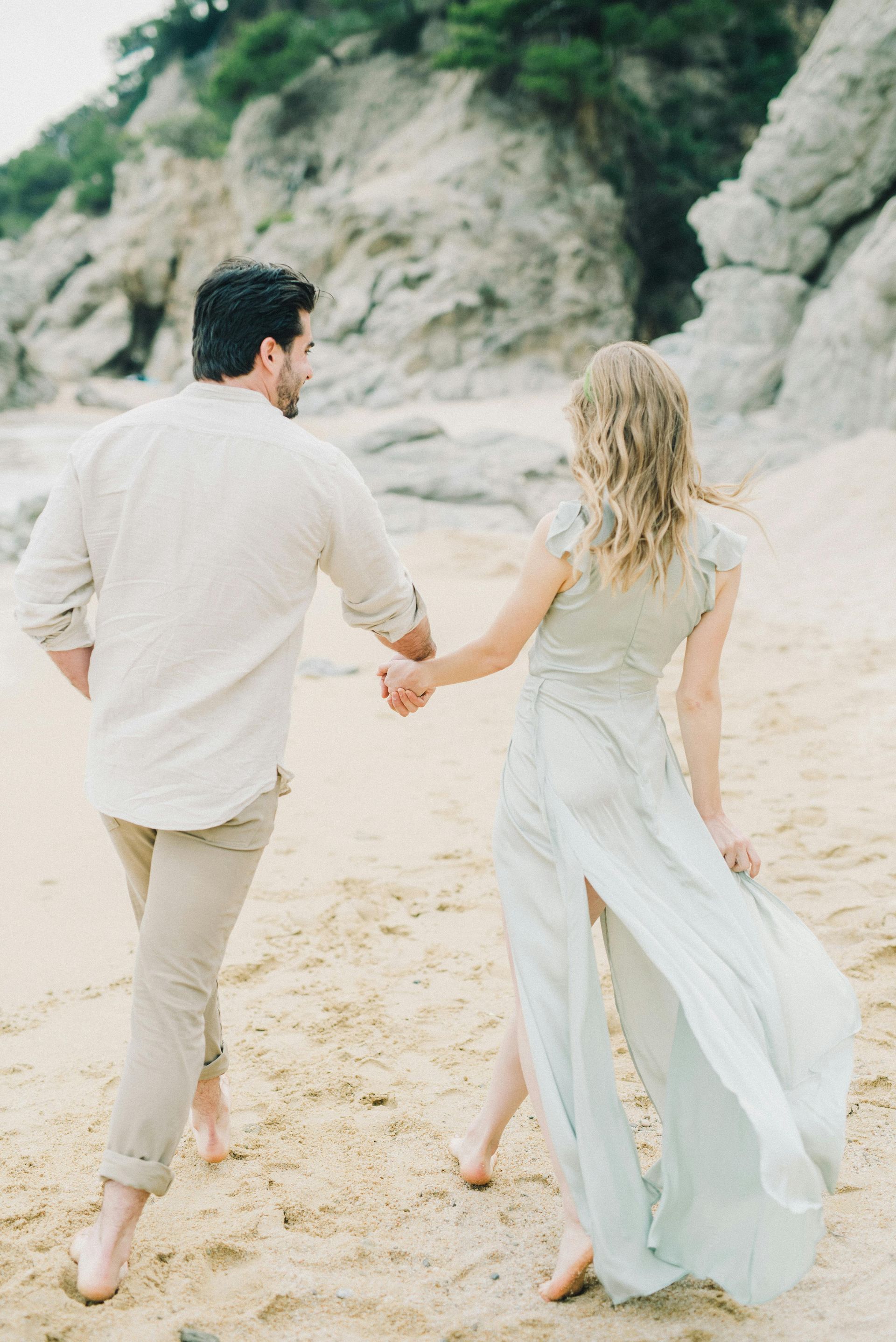 Couple holding hands running on sandy beach; man in beige, woman in light blue dress.