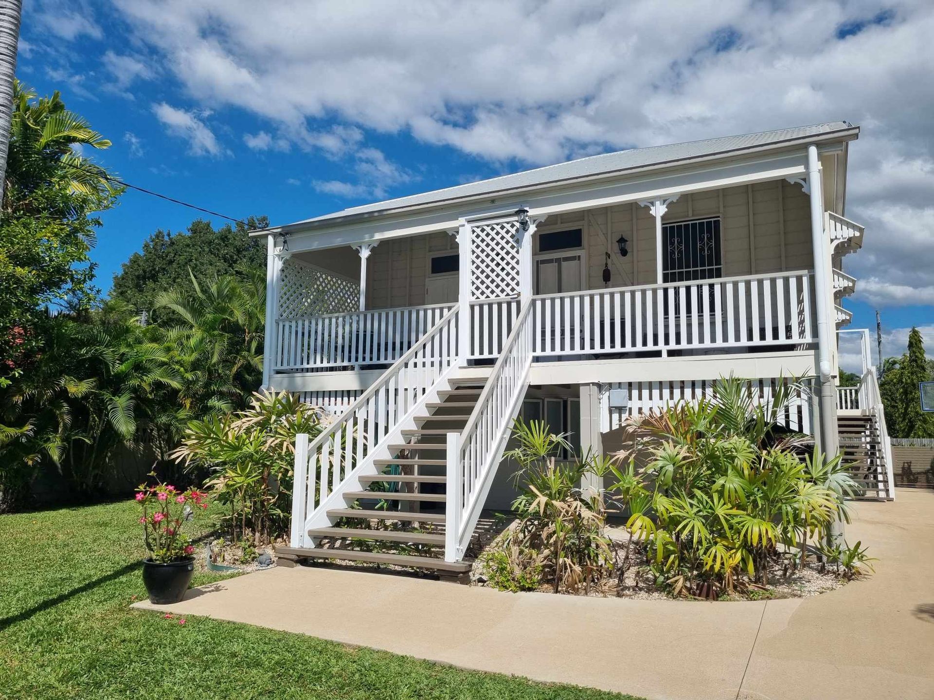 Two-story white house with wooden stairs and porch, surrounded by lush green plants under a blue sky.— Israel Jones Painting in Vincent, QLD
