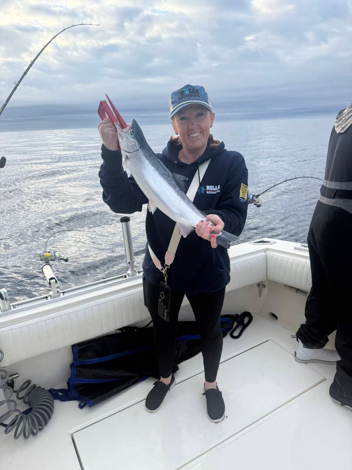 Woman on a boat holding a silvery fish with a red lure, smiling. Ocean background.