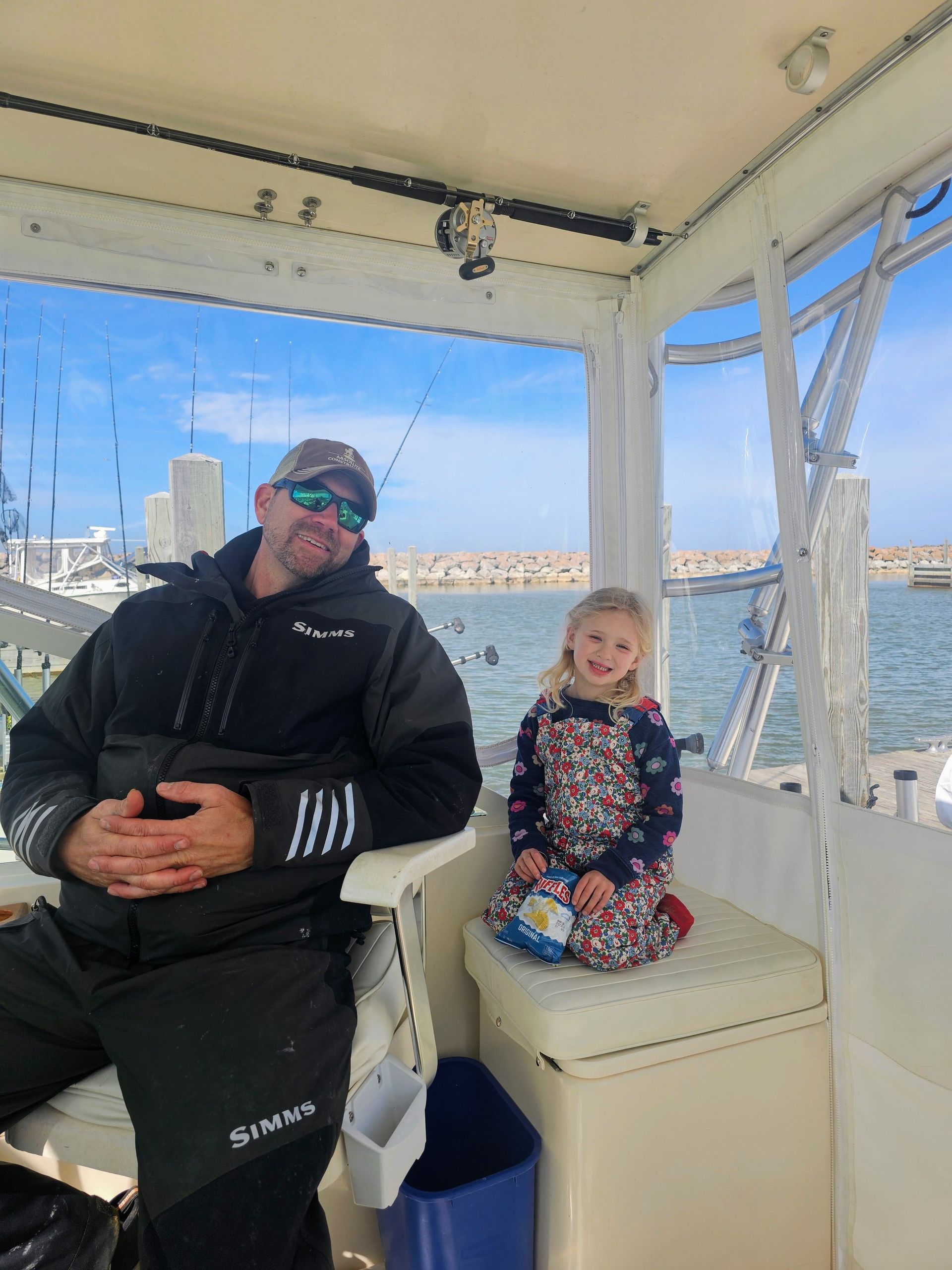 Man and child on a boat. Man in black jacket, child in floral dress. Smiling, sunny day with water view.