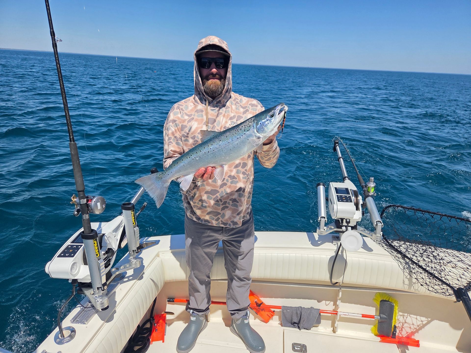 Man on a boat holds a large salmon, smiling. Sunny day, open water, fishing rods visible.