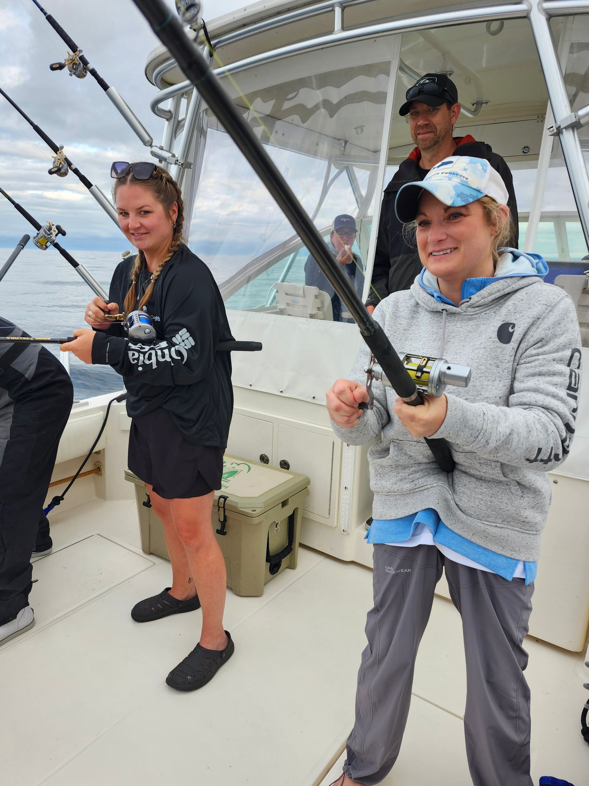 Two women and a man fishing on a boat, overcast sky, holding fishing rods, smiles.