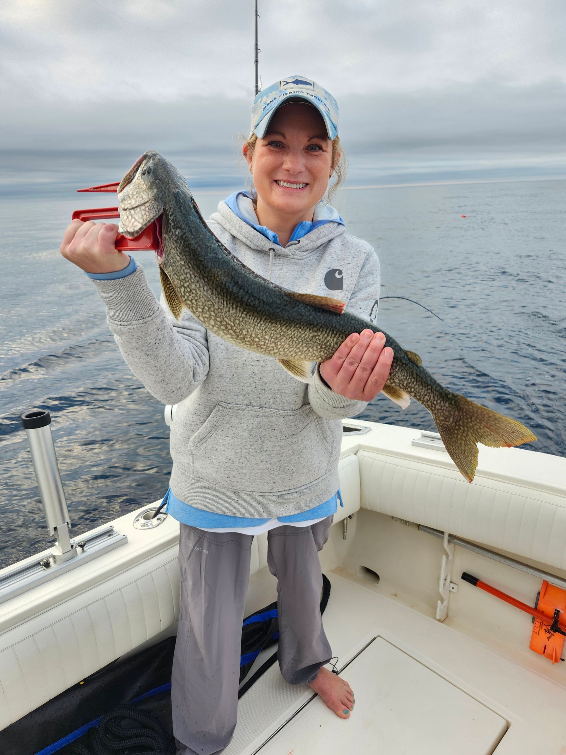 Woman on a boat holding a fish; she smiles. Gray sweater, gray pants, blue and gray hat; outdoors, lake.