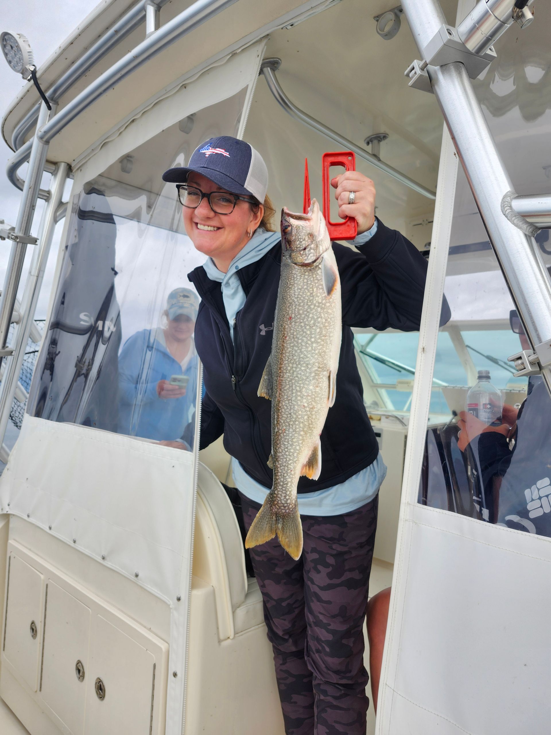 Woman on a boat, smiles, holding up a fish with a scale. Another person visible.