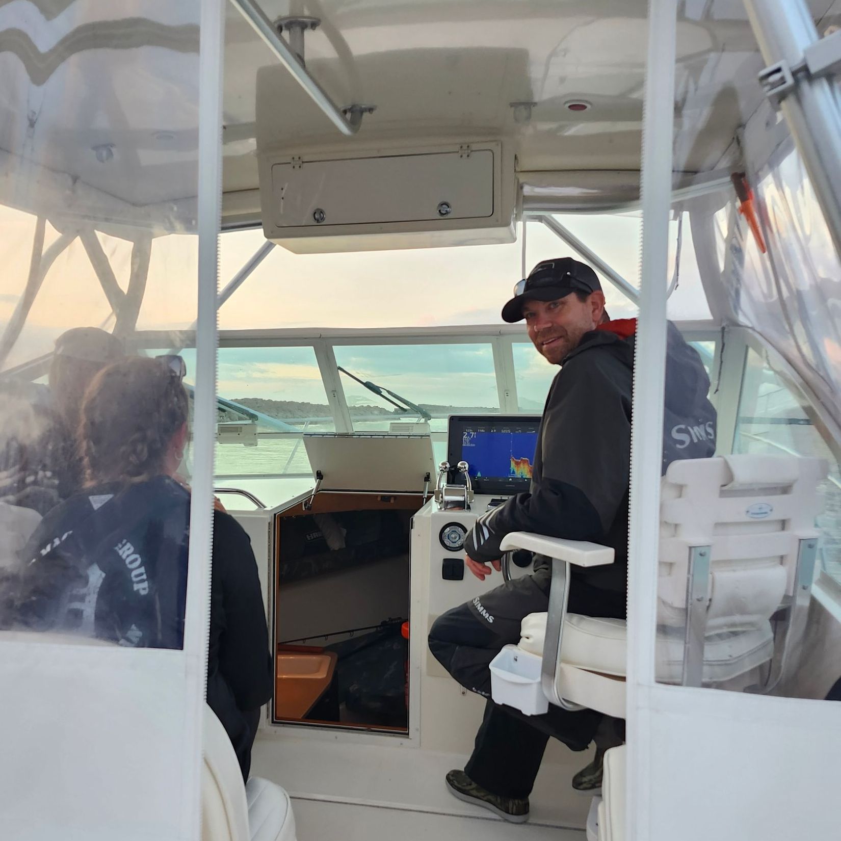 Man smiles driving a boat on a body of water. Passengers in the background.