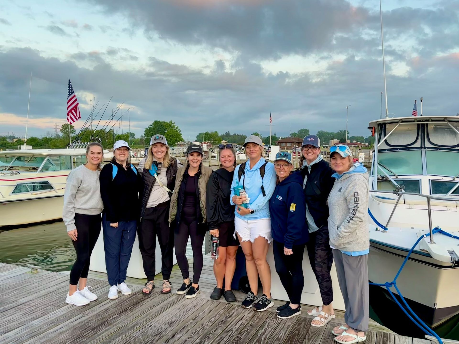 Group of women on a dock near boats, cloudy sky. Most wear caps and athletic wear. American flag visible.