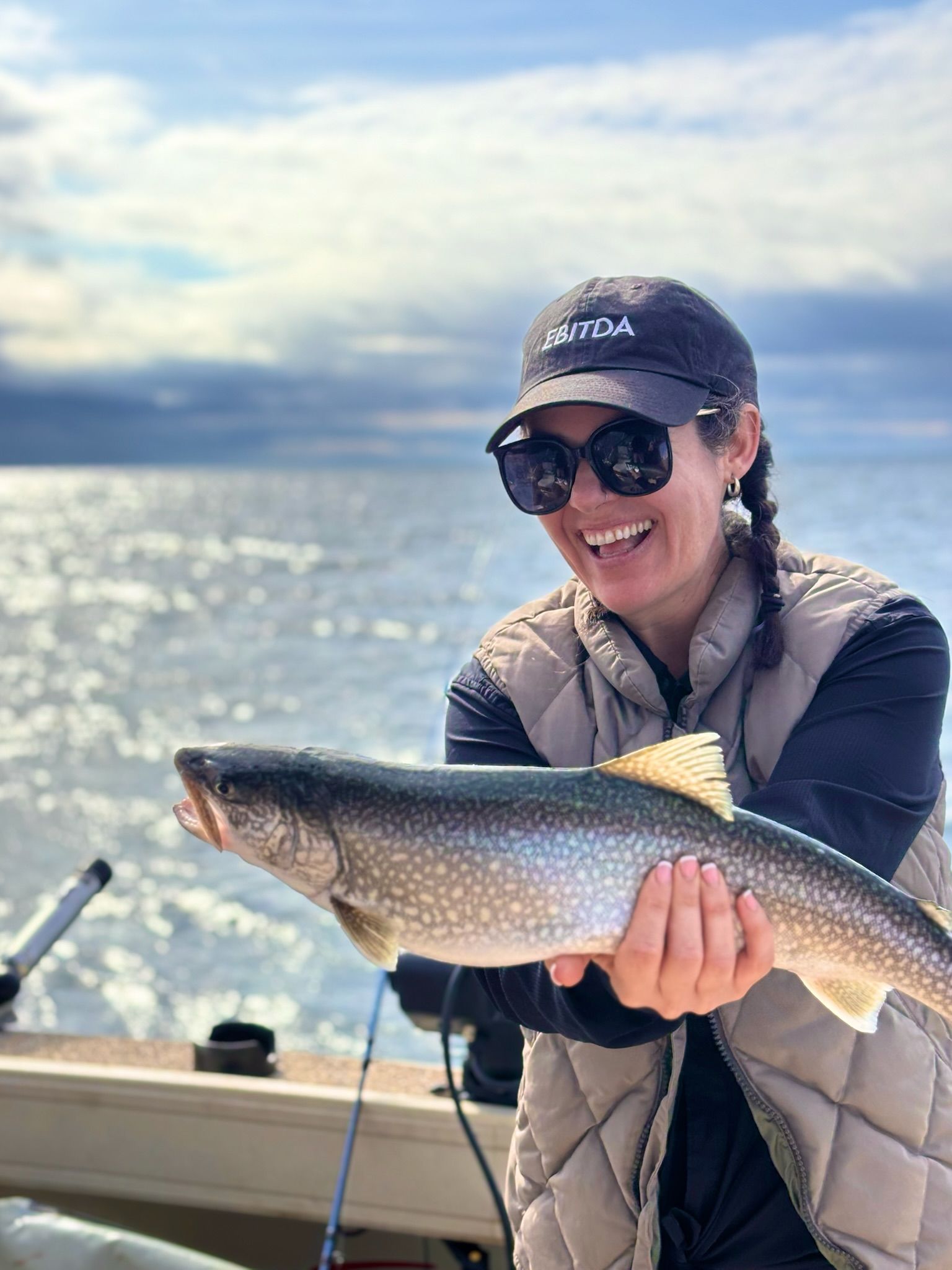 Woman smiling, holding a large fish on a boat. Wearing sunglasses, a hat, and vest; water and sky in the background.
