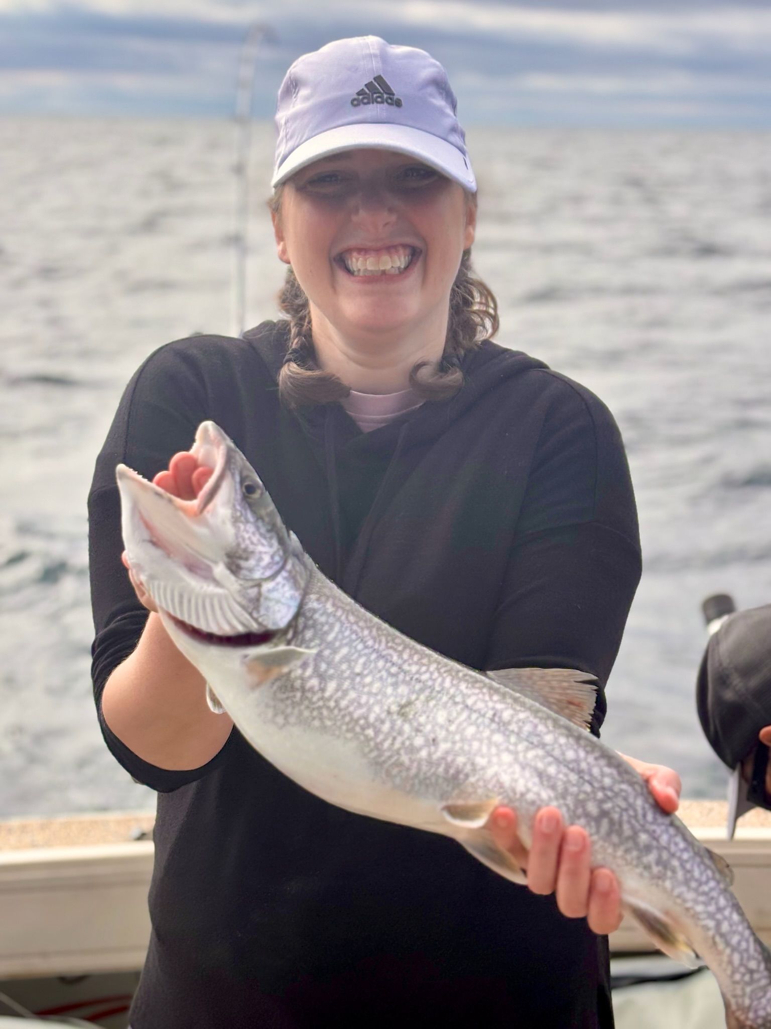 Woman smiling, holding a large fish with mouth open. On a boat, against a body of water.
