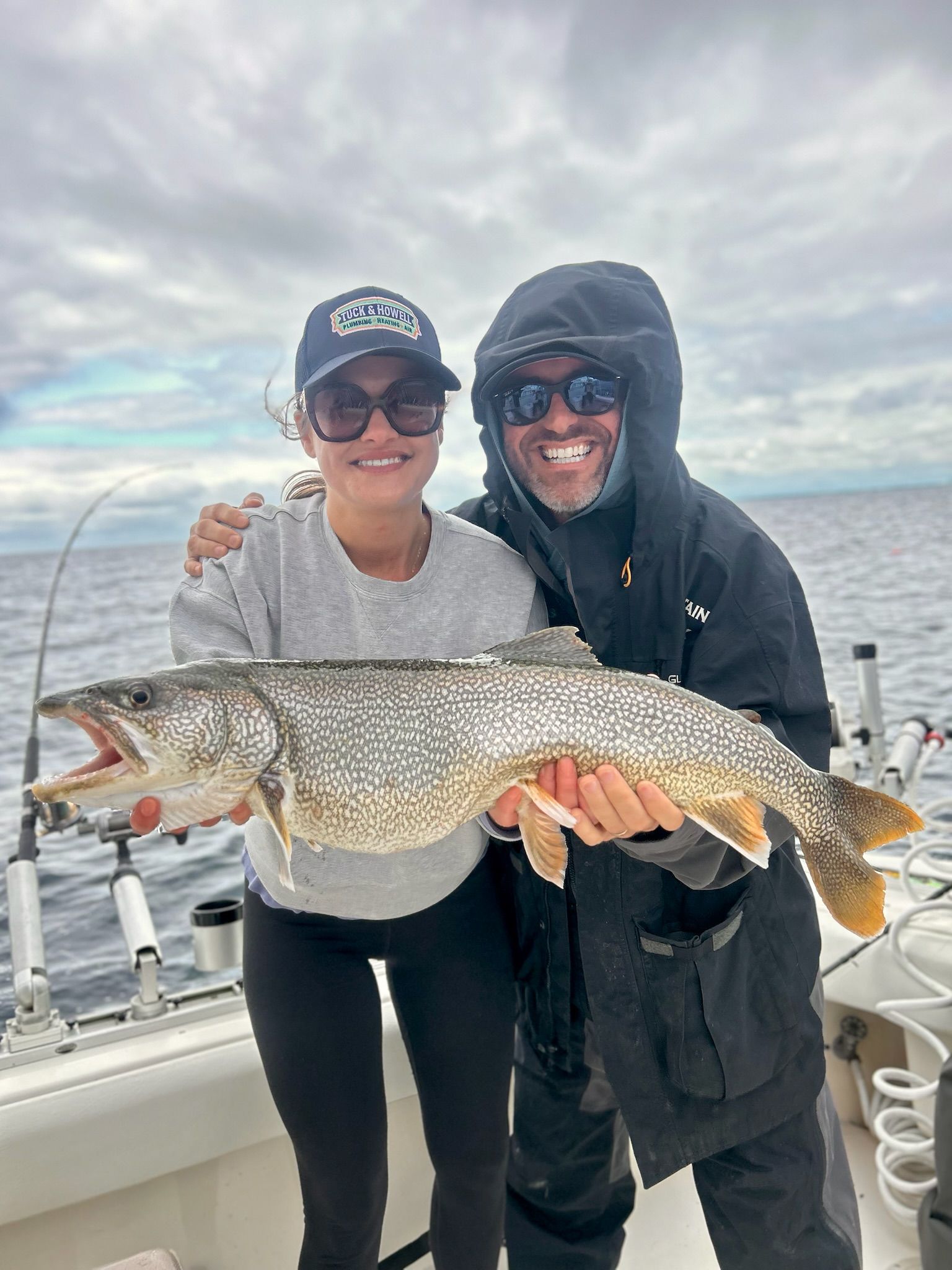 Couple on a boat smiling, holding a large spotted fish they caught, cloudy sky.