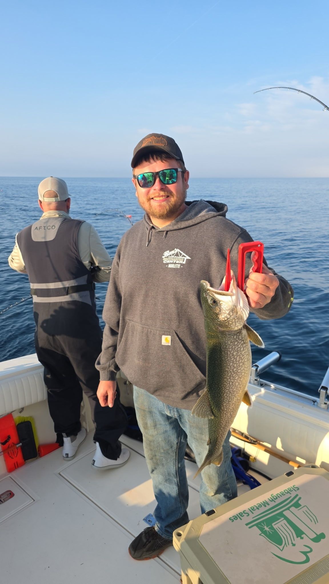 Man smiling, holding a large fish on a boat, another man fishing in the background, sea.