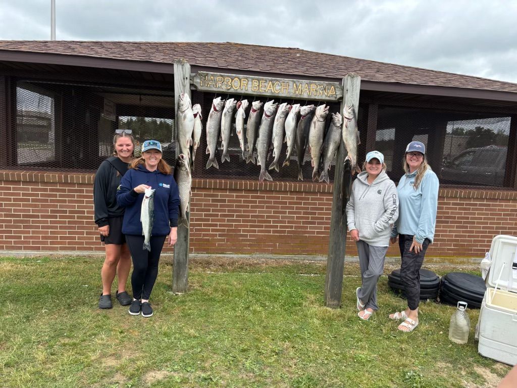 Four women stand with their catch; many fish hang from a wooden rack outside a brick building, overcast day.