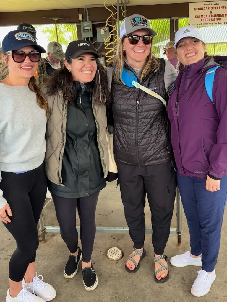 Four women smiling, posing closely together outside; one wears a vest and cap, others hats and jackets.