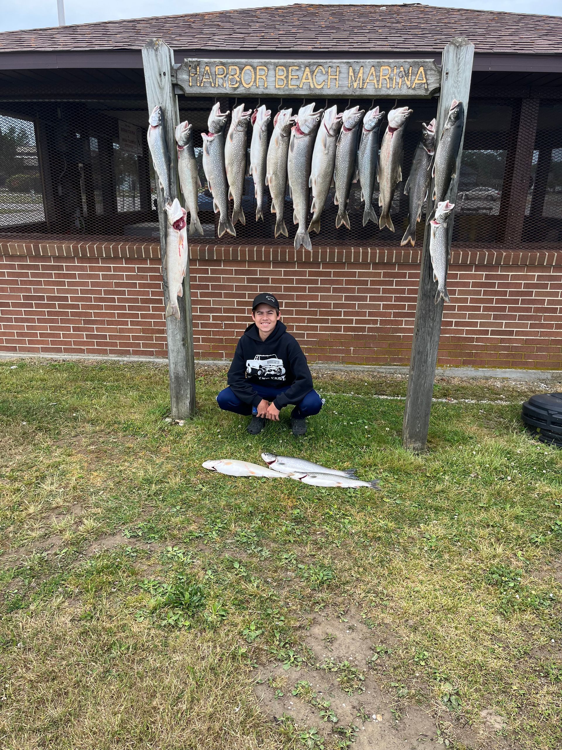 Young person crouches in front of a rack of fish, next to a brick building. They are outside on grass.