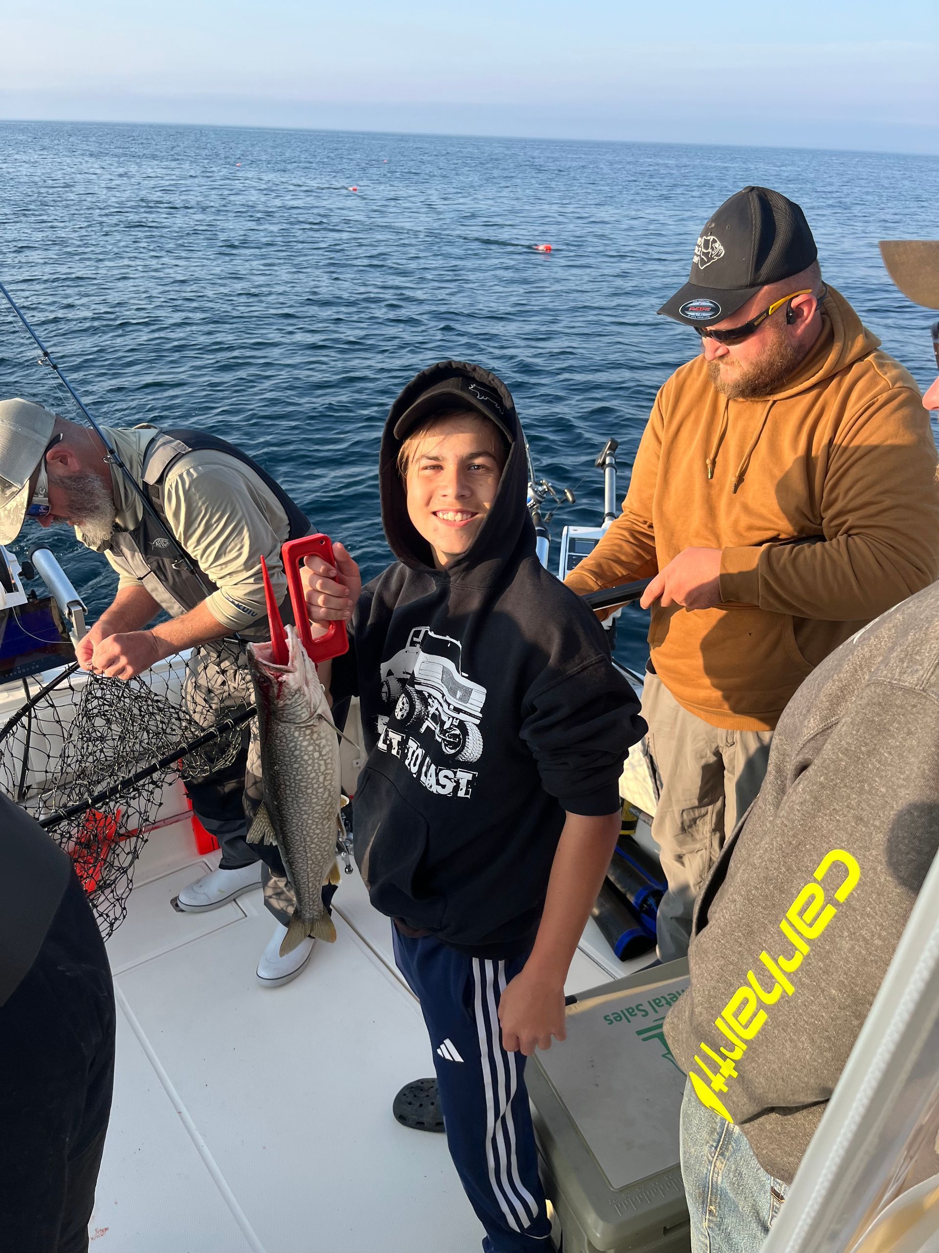 A young boy on a boat holds up a fish, smiling. Two men stand nearby. Ocean in background.