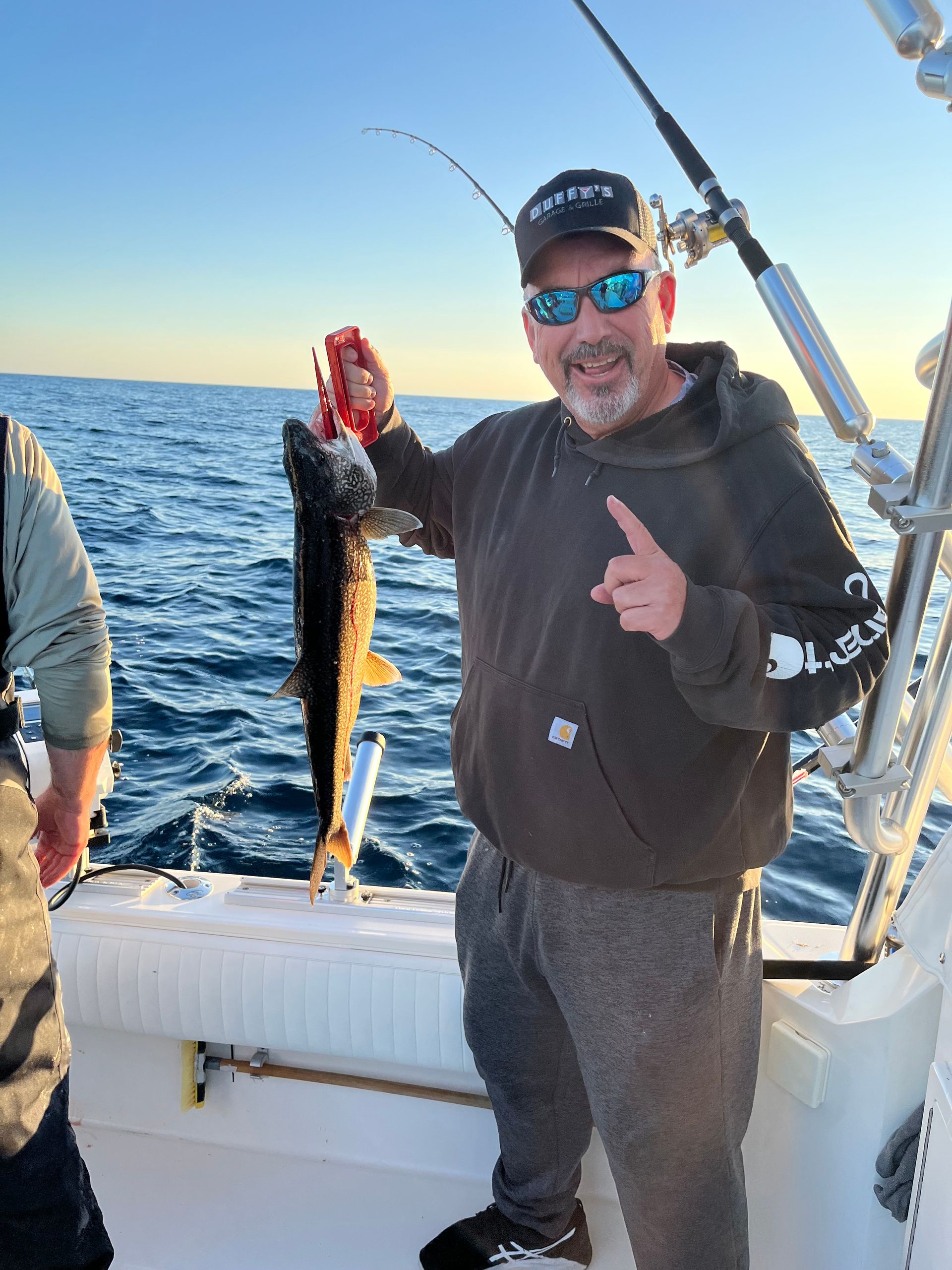Man on a boat holding a fish; points at it, smiling. Blue water and sky.