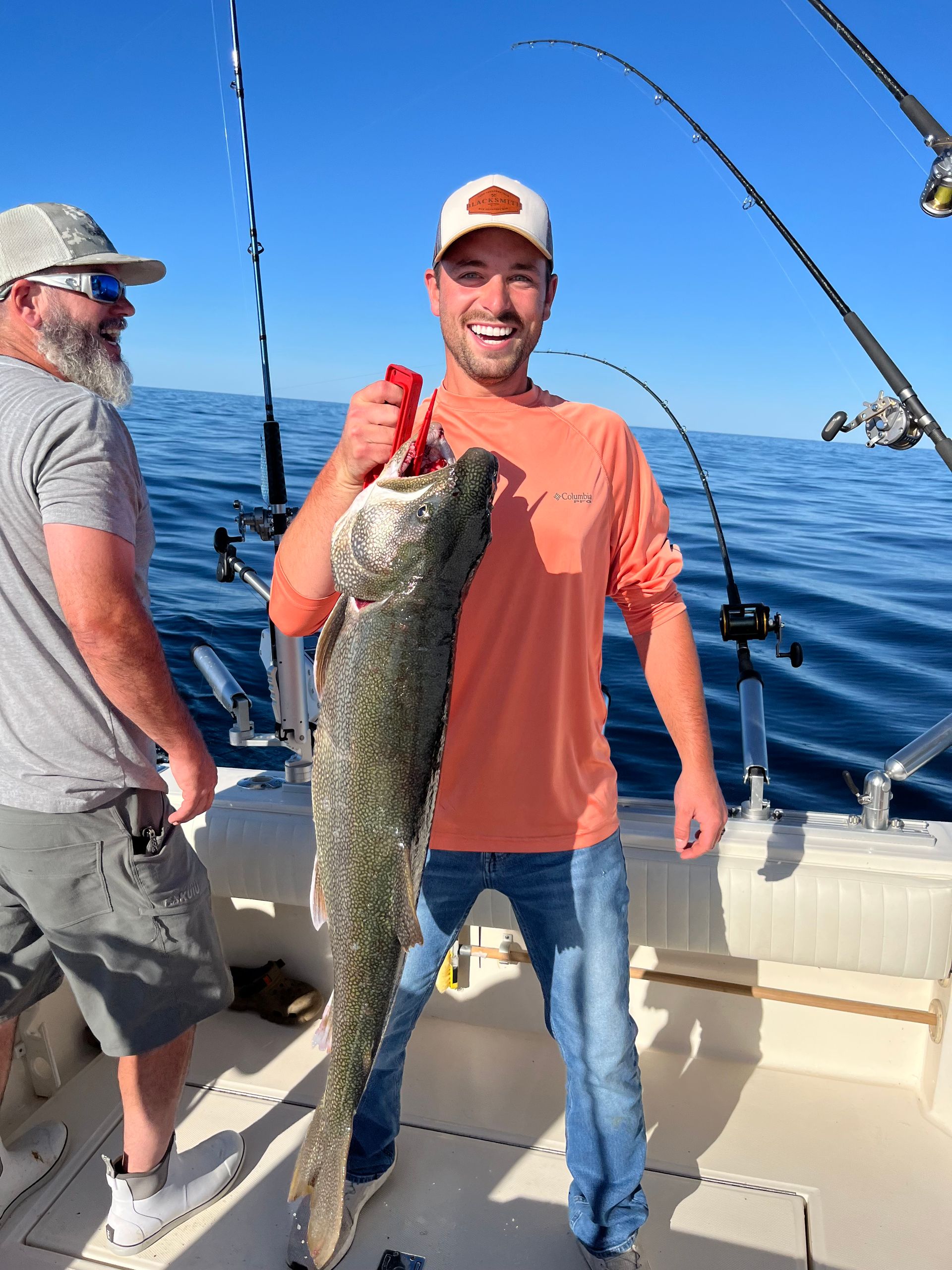 Man on a boat holding a large fish, smiling. Another man stands beside them with fishing poles. Outdoors, blue water.