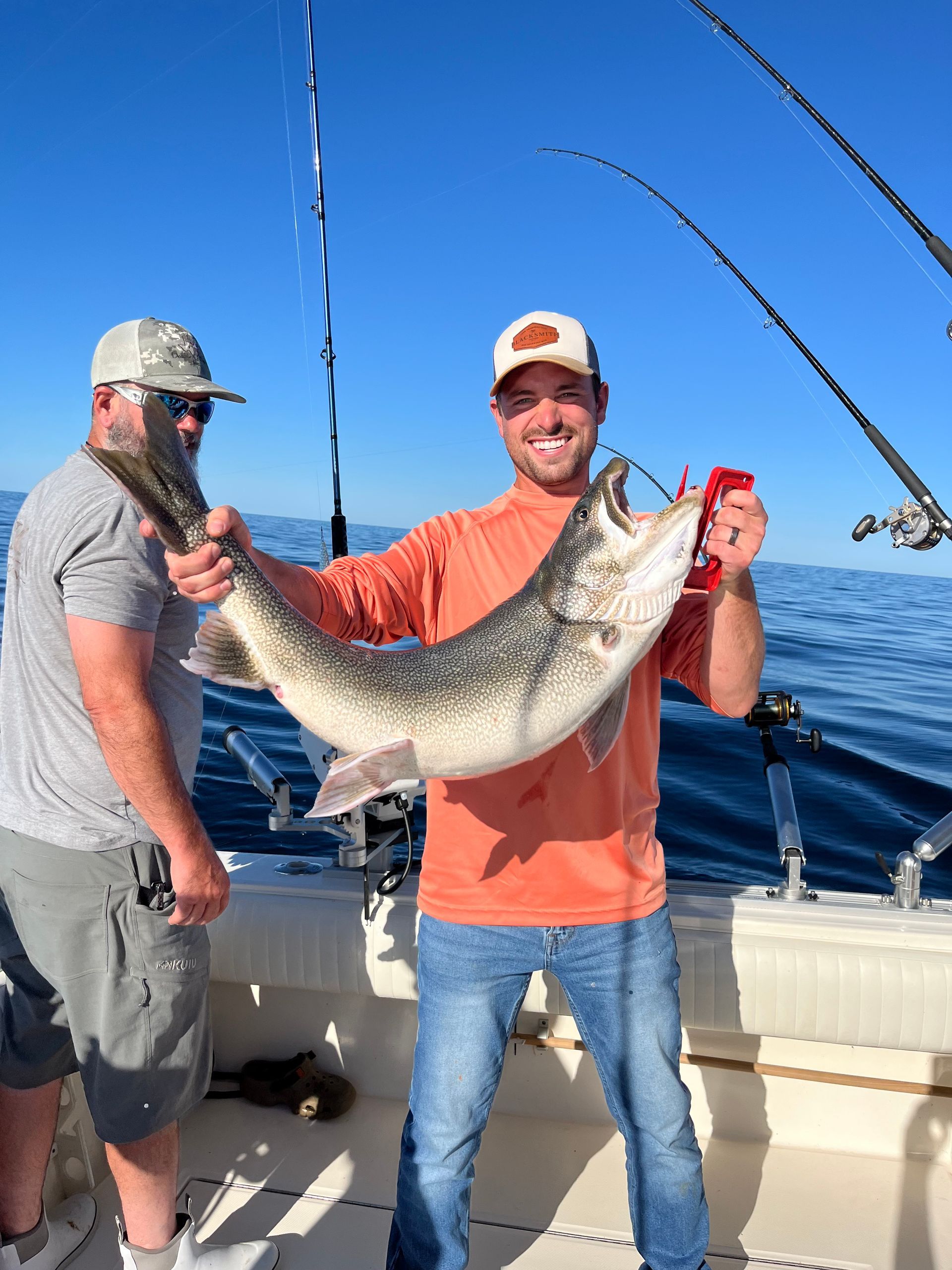 Two men on a boat hold up a large fish they caught; sunny day, blue water, one smiles.