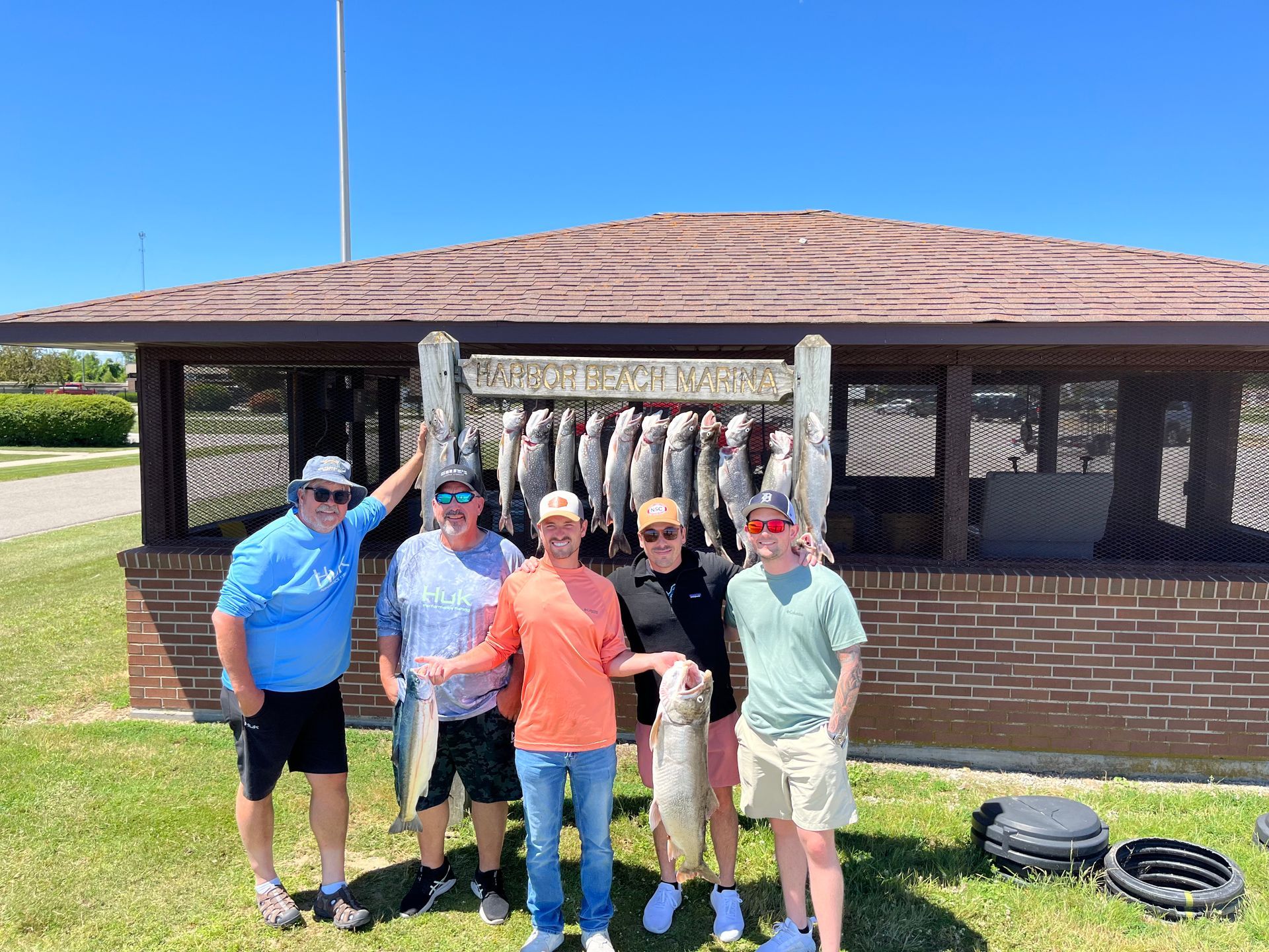 Group of men with fish they caught at a lakeside pavilion on a sunny day.
