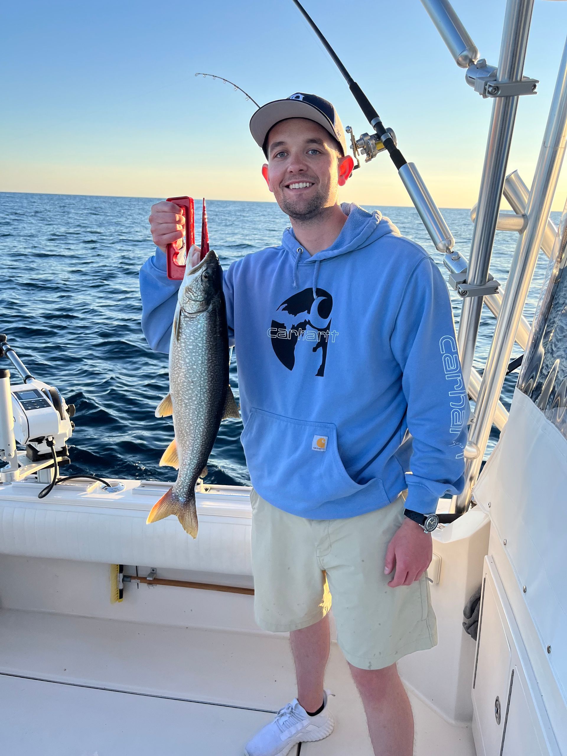 Man on boat holding fish, smiling. Wearing blue hoodie, tan shorts. Holding can; ocean background.