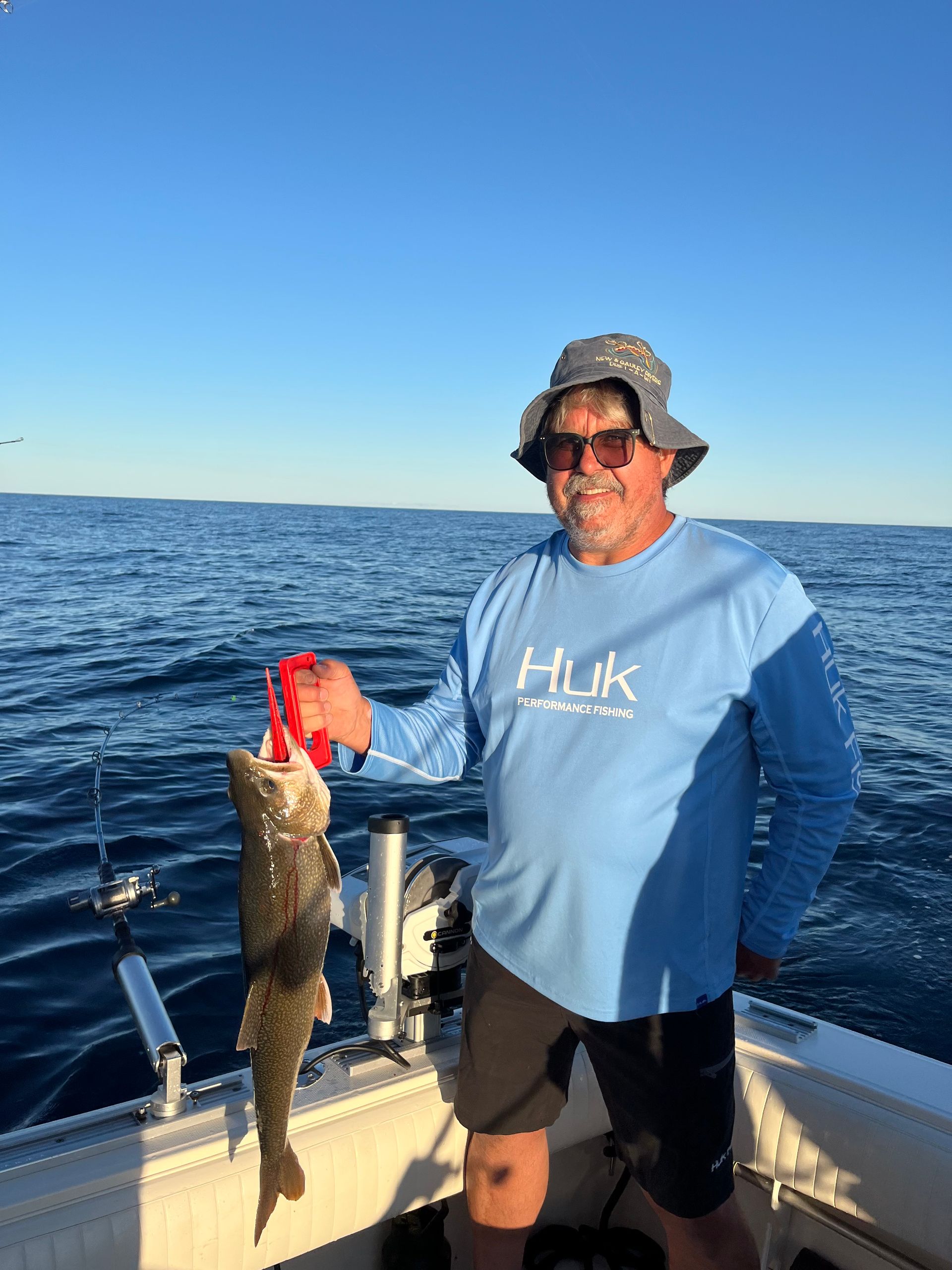 Man on a boat holds up a fish, blue water and sky in the background. He wears sunglasses, a hat, and a blue shirt.